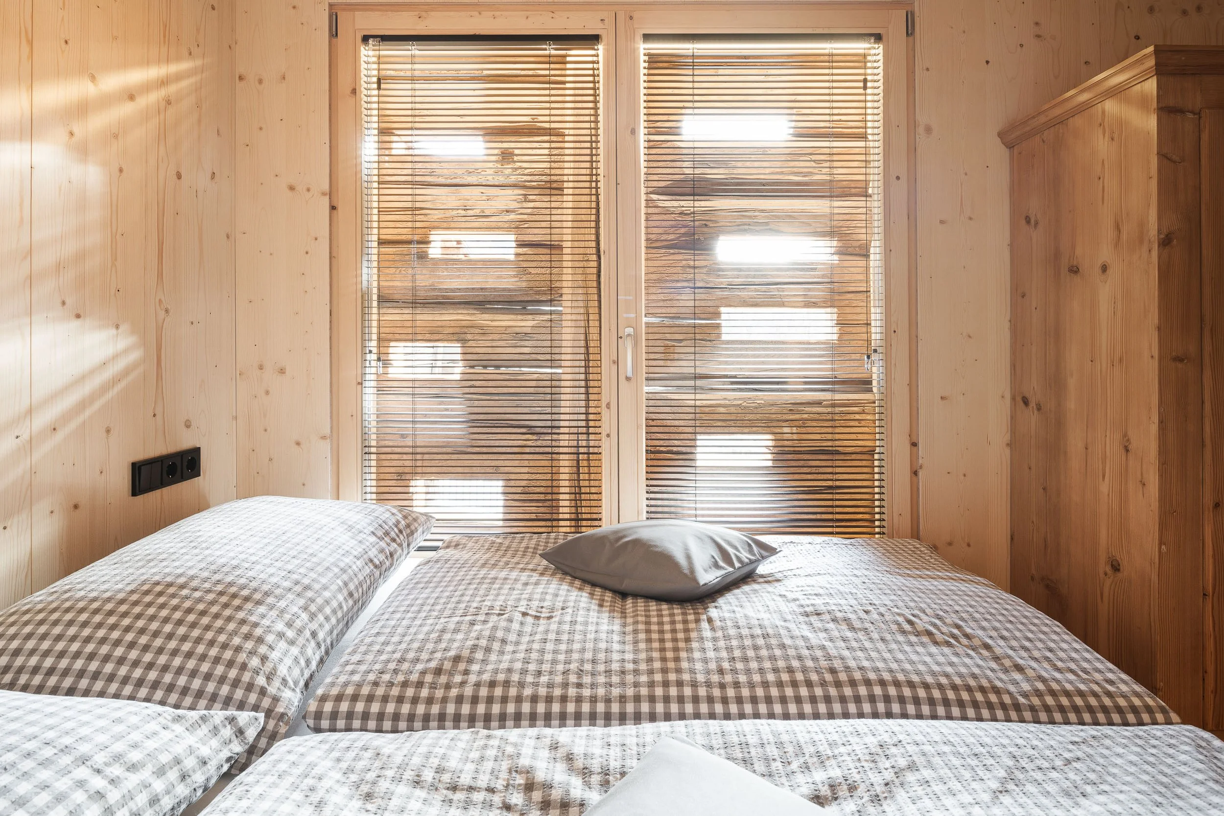 Ein Schlafzimmer mit Holzwänden, zwei Betten mit karierten Bettzeug und einem Kissen, einen großen Fenster mit Holzbalkenjalousien.