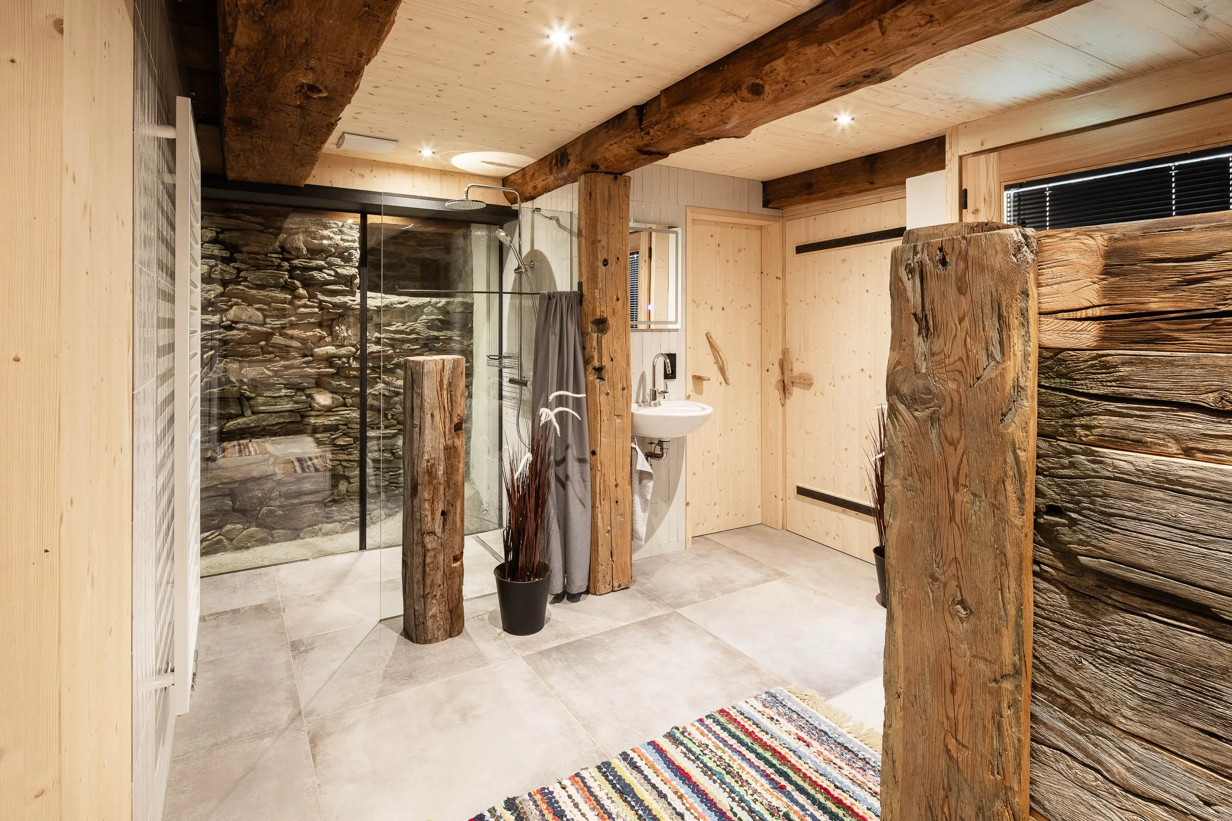 Ein Badezimmer mit Holz- und Naturstein-Design, Holzbalken an der Decke, eine begehbare Dusche mit Steinen im Hintergrund, Waschbecken, buntem Teppich, schwarzer Fensterjalousie und Pflanzen in schwarzen Töpfen.