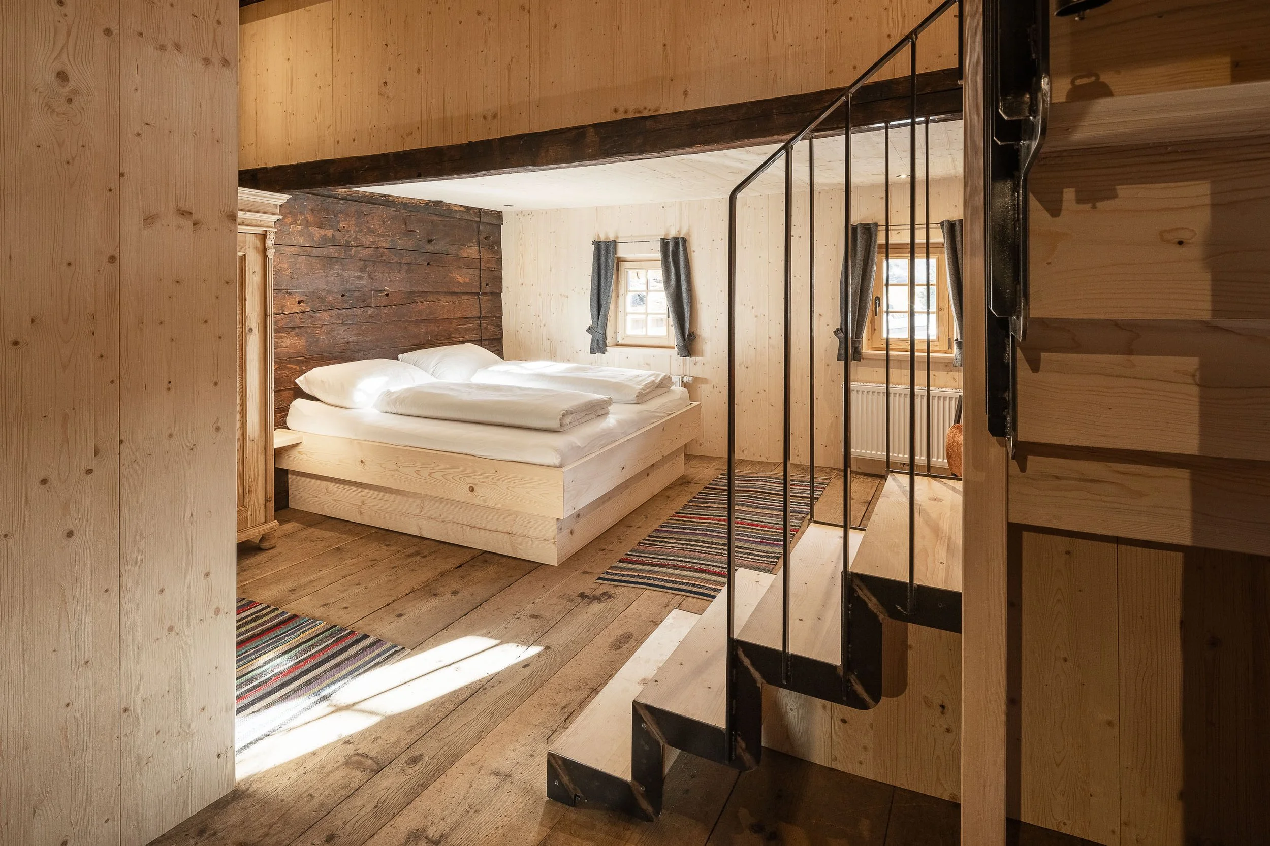 Ein Schlafzimmer mit Holzdecke, Holzwänden und Holzboden. Es gibt ein Doppelbett mit weißen Bettwäsche, zwei Fenster mit grauen Vorhängen, und eine schwarze Treppe führt nach oben.