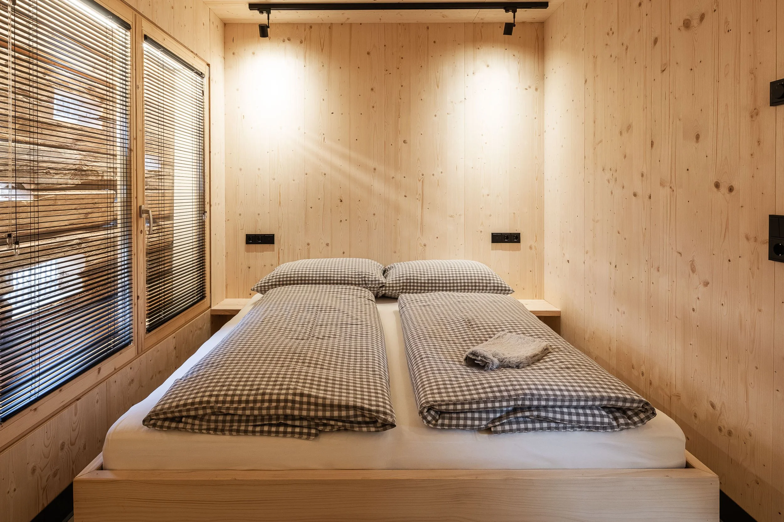 Ein Schlafzimmer mit Holzwänden und -decken, zwei Betten mit karierten Bettwäsche, eine abgelegte Decke, Fenster mit Holzjalousien und elektrische Steckdosen an der Wand.