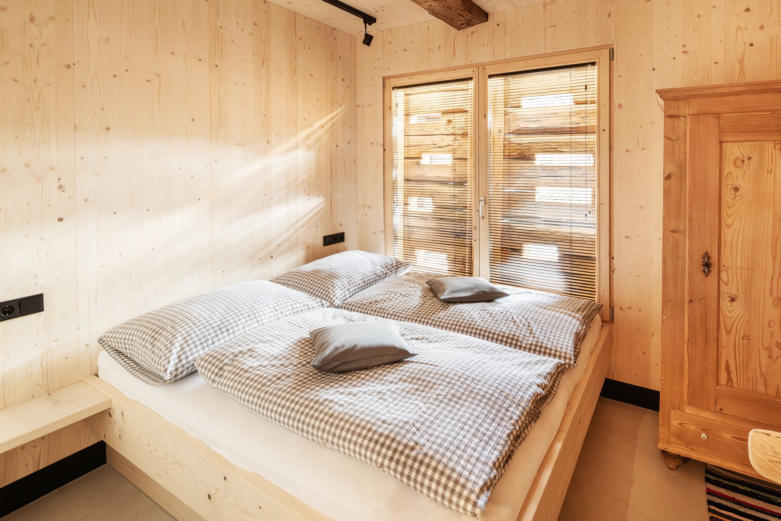 Schlafzimmer mit Holzverkleidung, Doppelbett mit karierten Bettwäsche, Fenster mit verstellbaren Jalousien, Holzschrank, Nachttisch und Teppich.