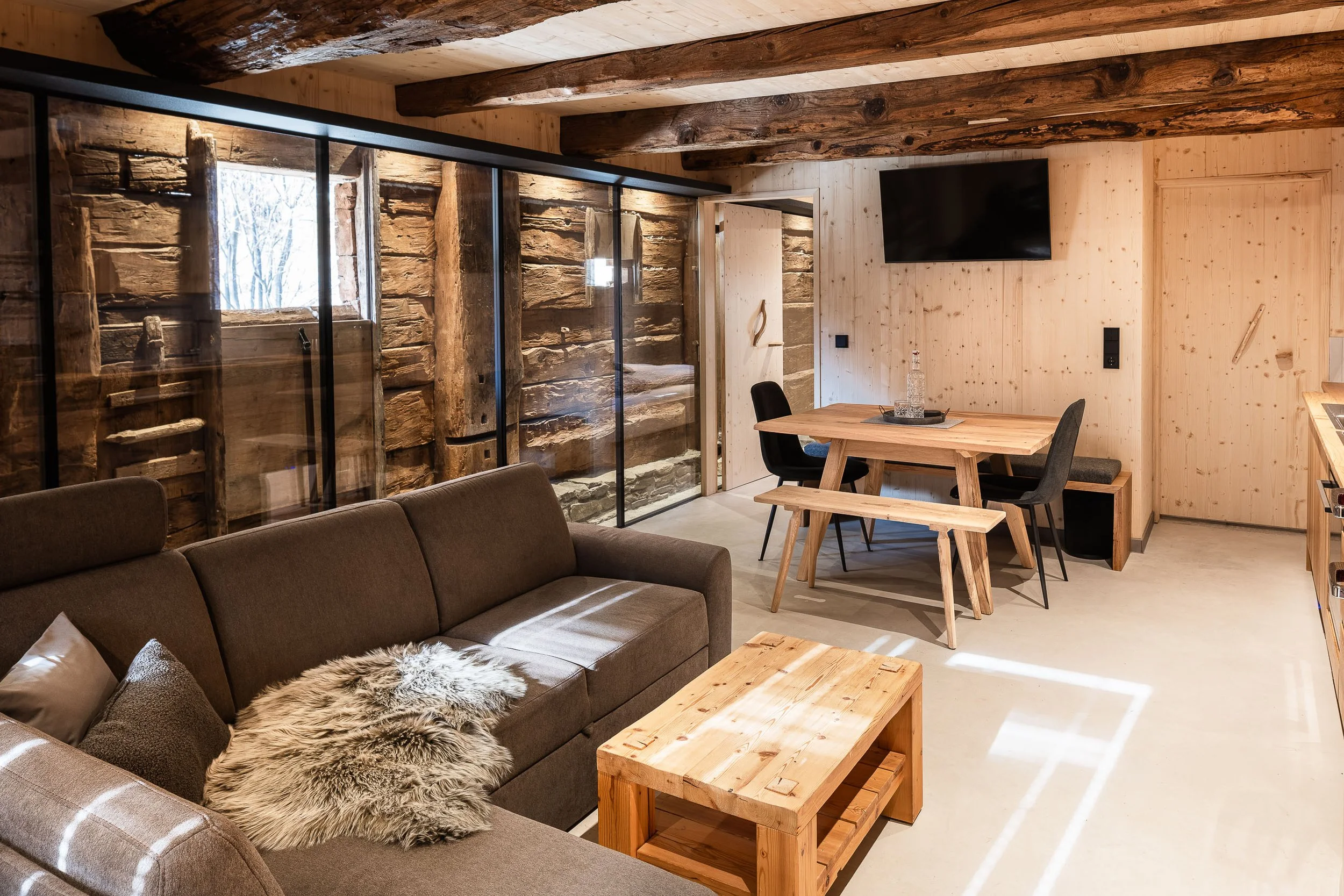Gemütliches Wohnzimmer mit Holzwänden und Decke, einem braunen Sofa mit Fell- und Kissen, einem Holztisch, Esstisch mit Stühlen, Fernseher an der Wand, große Fenster mit Blick nach draußen.