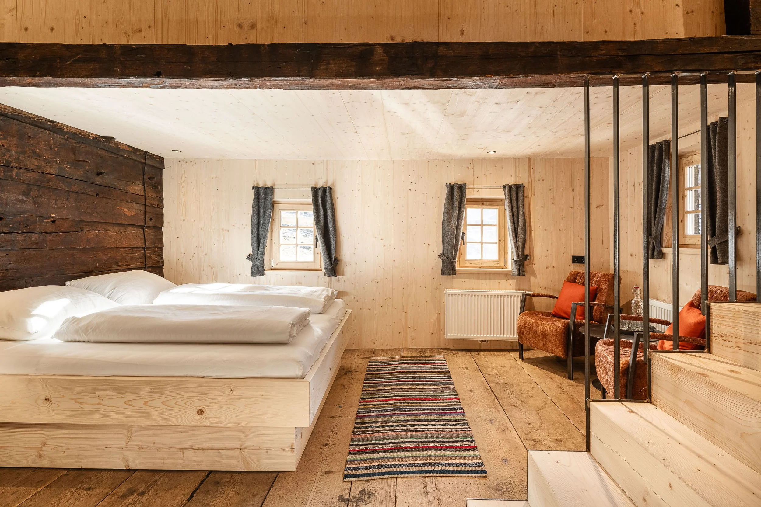 Innenaufnahme eines Holzhauses mit Doppelbett, mehreren Fenstern mit Vorhängen, einem Teppich und Sitzgelegenheiten.