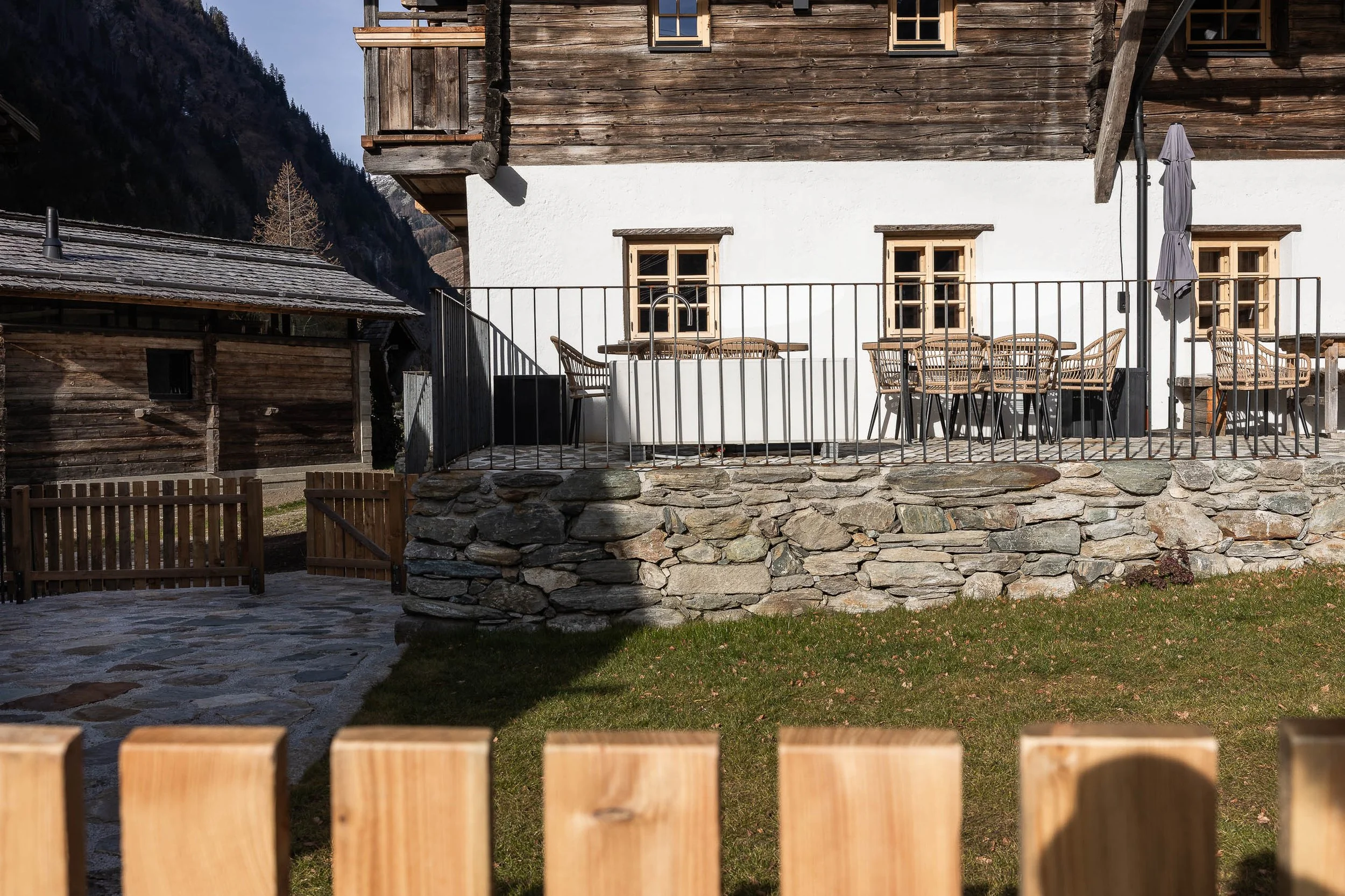 Ein Haus im alpinen Stil mit einer Terrasse, Holzmöbeln und einer parapetförmigen Brüstung aus Metall. Das Haus hat Holzwände oben und eine weiße Wand unten mit mehreren Fenstern. Es gibt einen Garten mit grünem Rasen und einen Steinweg, der zur Terrasse führt.