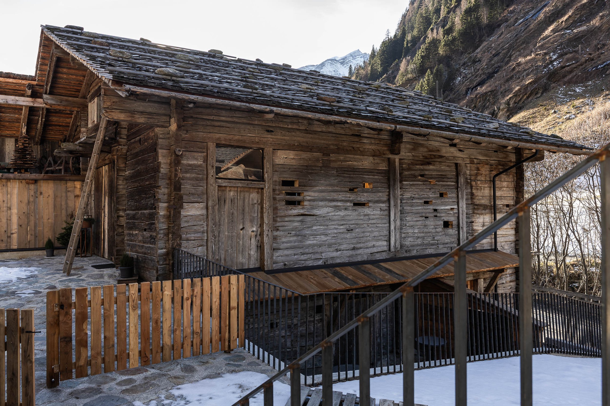 Ein holzgestricktes Haus in einer Bergregion mit verschneitem Boden, umgeben von Felswänden und Tannbäumen.