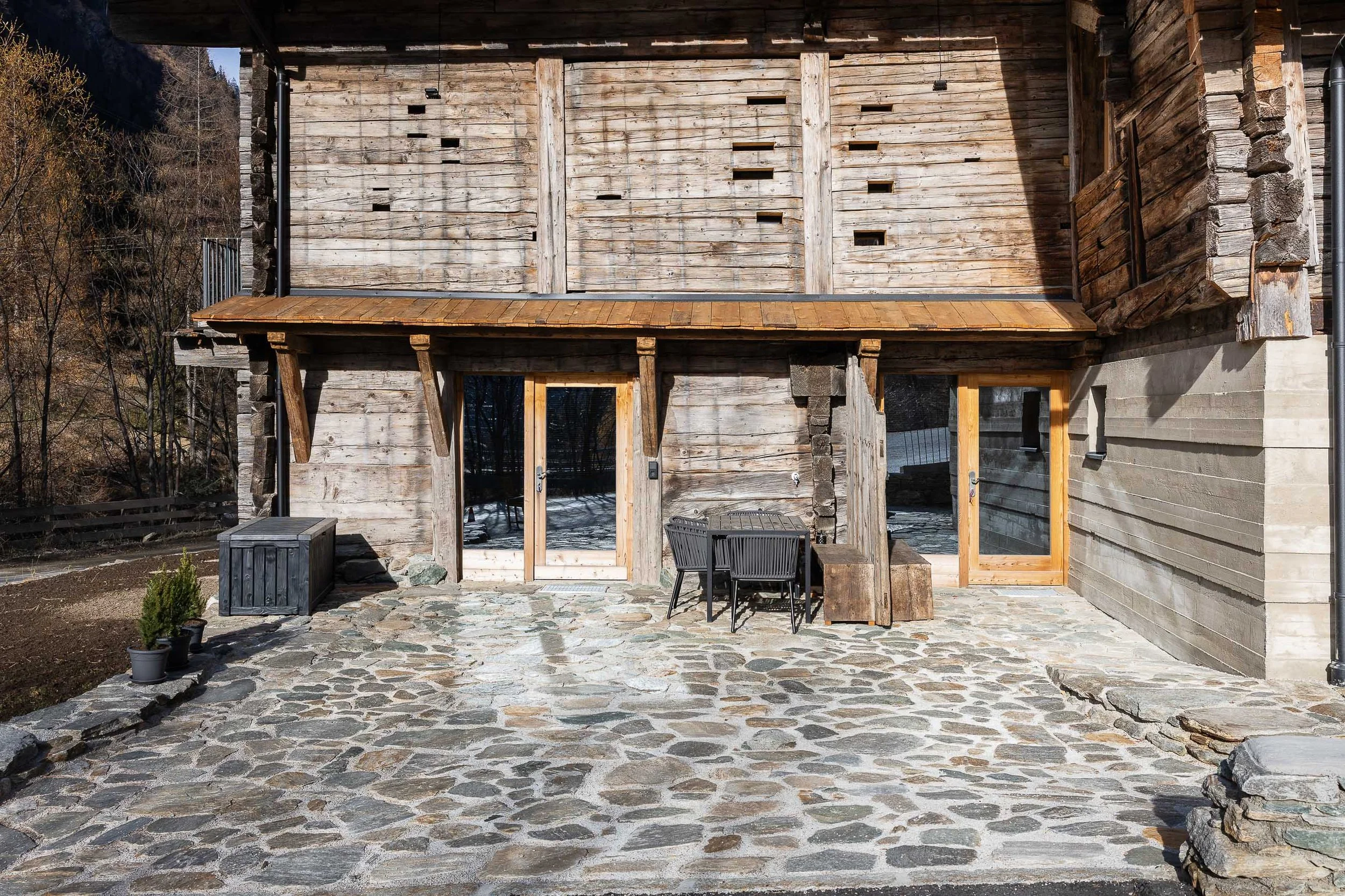 Holzhaus mit Terrasse und Pflanzkübeln vor einem Steinboden. Im Hintergrund sind Bäume und Berge sichtbar.