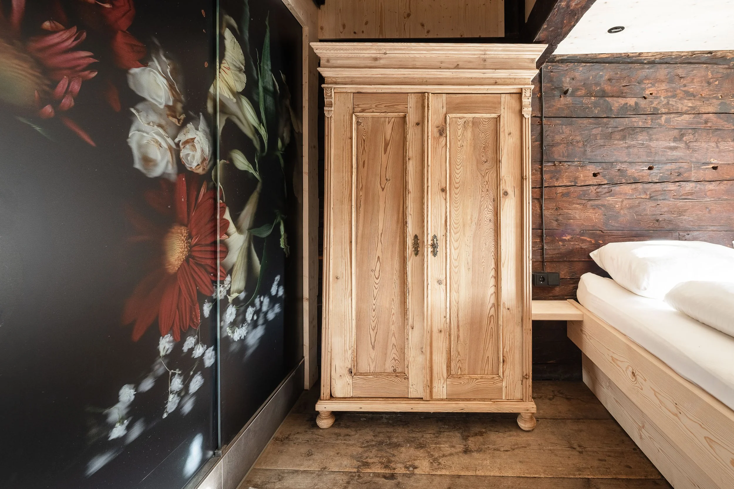 Ein Schlafzimmer mit Holzankleide, Holzlaminatboden, und Wandverkleidung aus Holzbrettern. Es gibt einen Bettrahmen mit weißen Kissen und eine dunkel gestaltete Wand mit Blumenmuster.