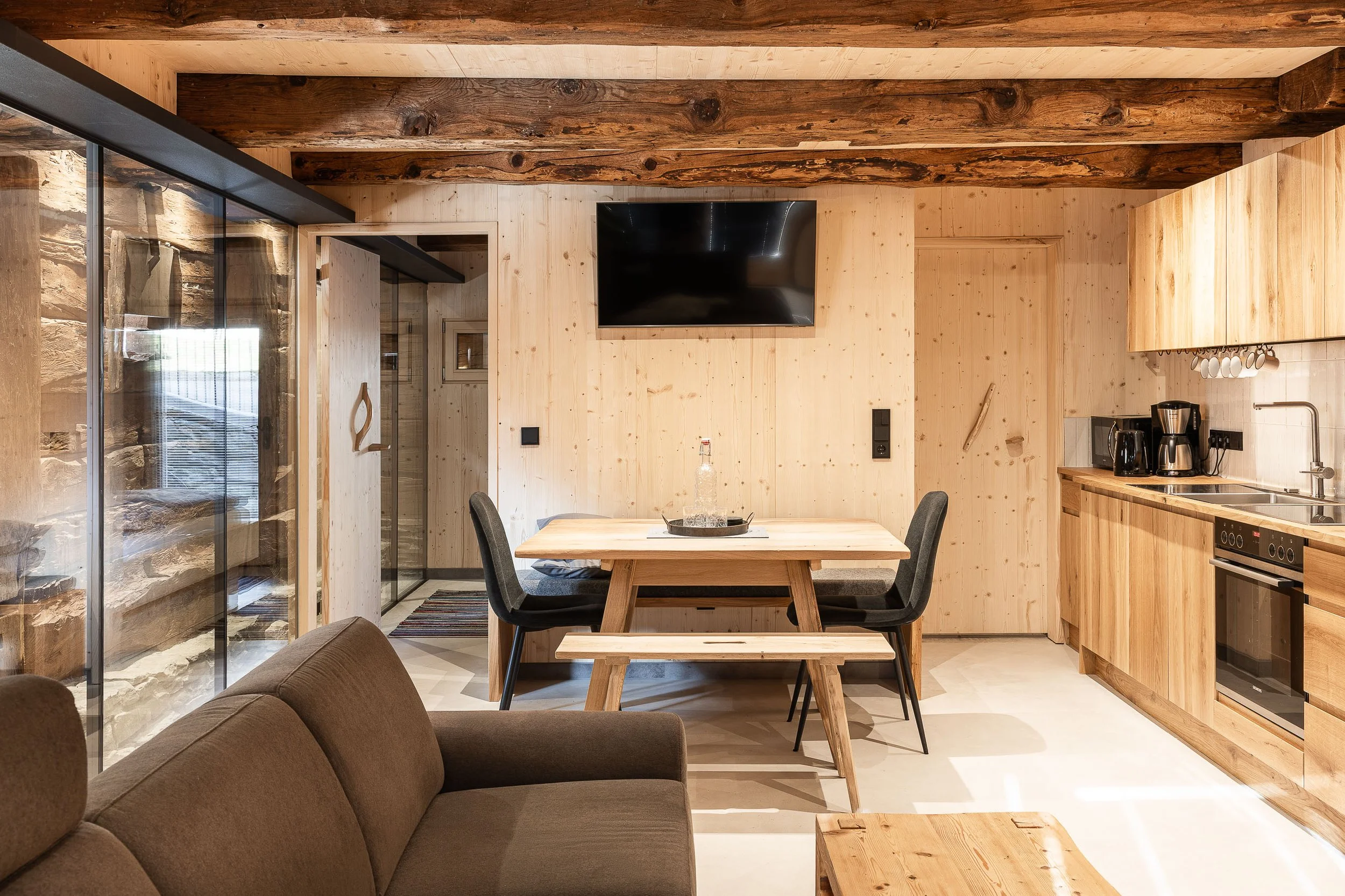 Wohnküche mit Holzvertäfelung, Esstisch, Fernseher an der Wand, Küche mit Mikrowelle, Kaffeemaschine und Herd, umliegende Holzwände und Deckenbalken.