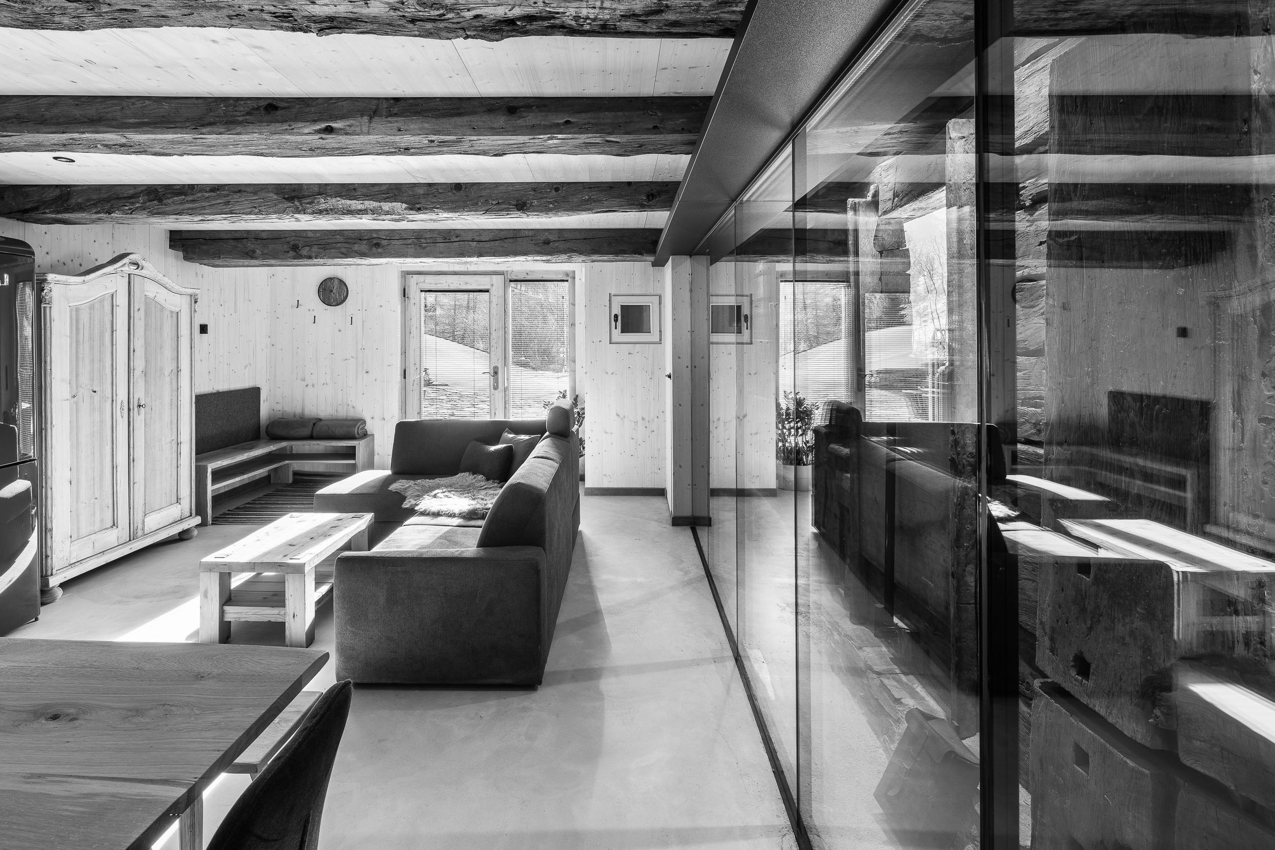 Innenansicht eines modernen Wohnzimmers mit Holzdeckenbalken, großen Fenstern und einem gemütlichen Sofa.