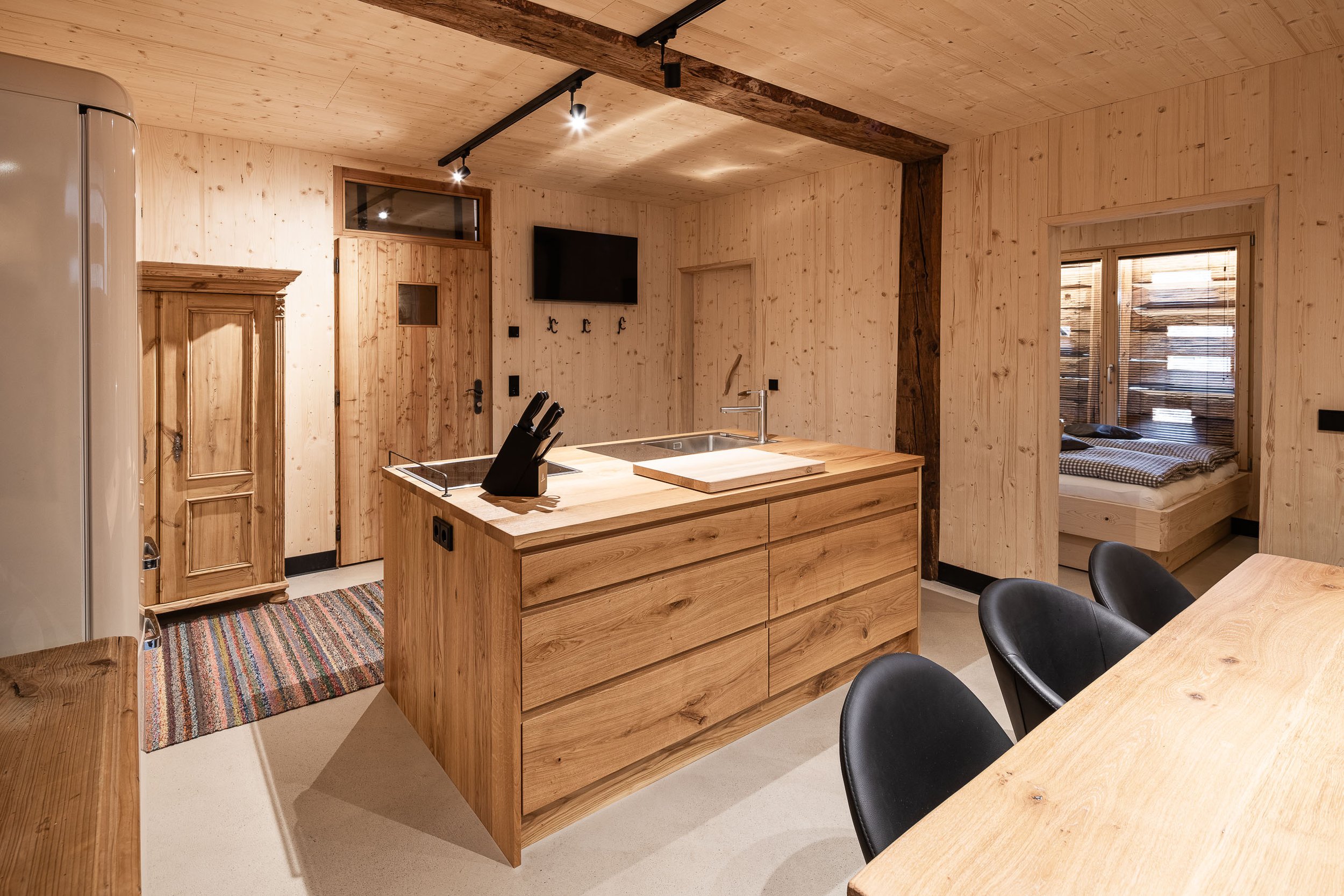 Innenraum einer holzvertäfelten Küche mit Kücheninsel, Esstisch und Stühlen, TV, Bett im Hintergrund, Fenster mit Jalousien, helles Holzdesign und Teppichboden.