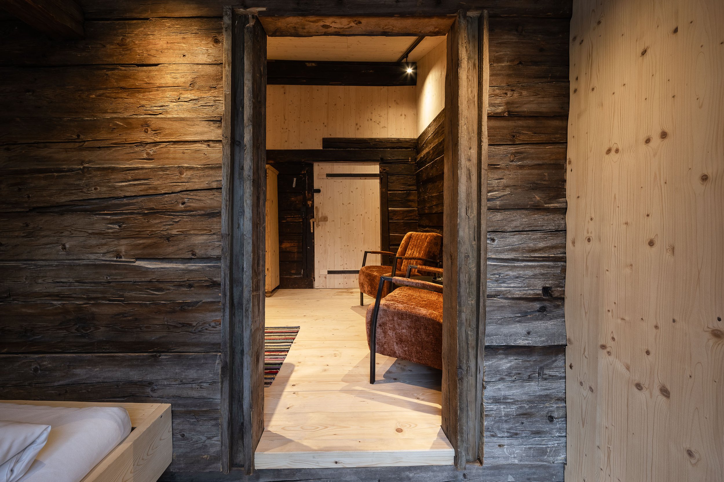 Innenaufnahme eines rustikalen Zimmers mit Holzwänden und -decken, Blick durch einen Türrahmen, sichtbar sind zwei Vintage-Stühle, eine farbige Vertikalarbeitsmatte auf dem Boden und ein Bett im unteren linken Bildbereich.