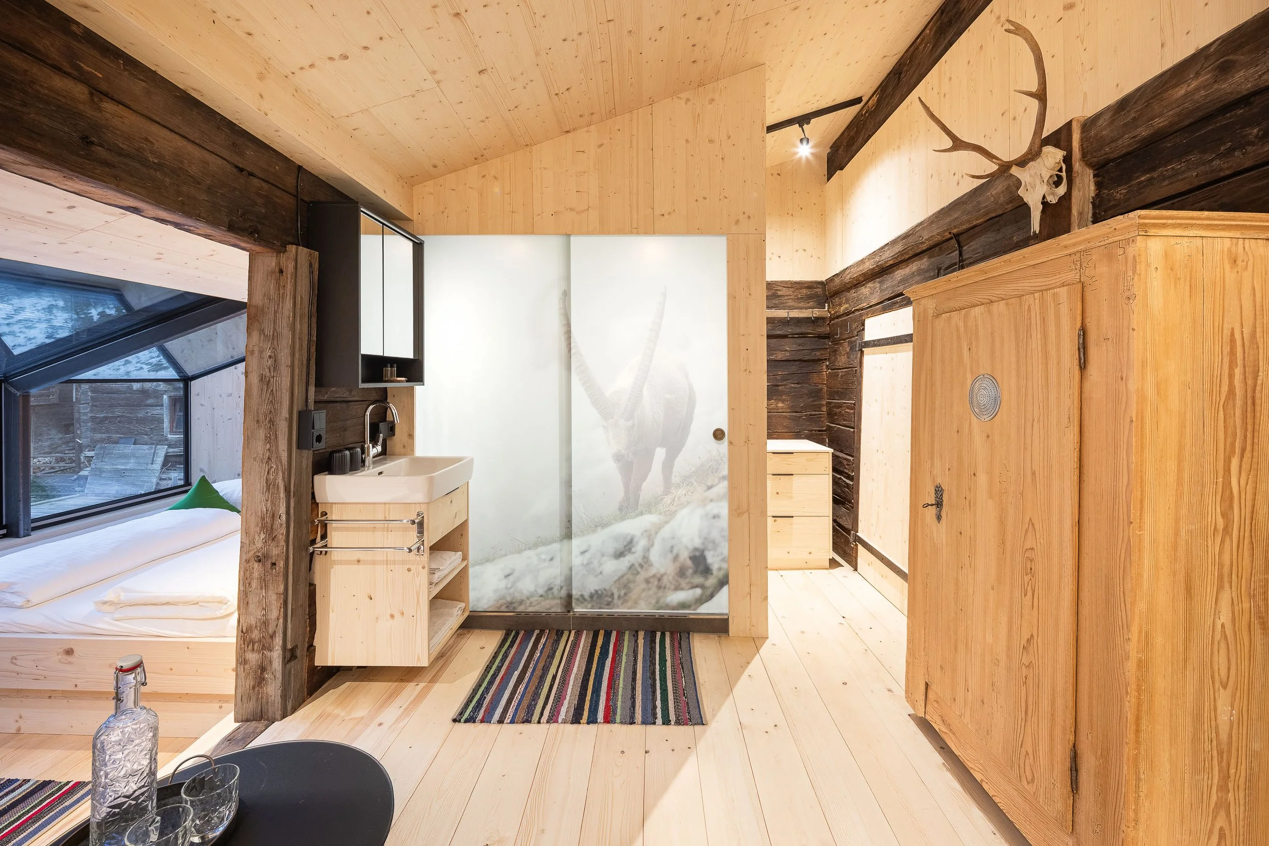 Innenansicht eines modernen Holzhotels mit Holzdecken und Holzwänden, einem Bett, einem Waschbecken, einem skulpturalen Wandbild eines Pferdes, einem Pumakopf mit Geweih an der Wand, einem Schrank und einer bunten Teppichstraße.