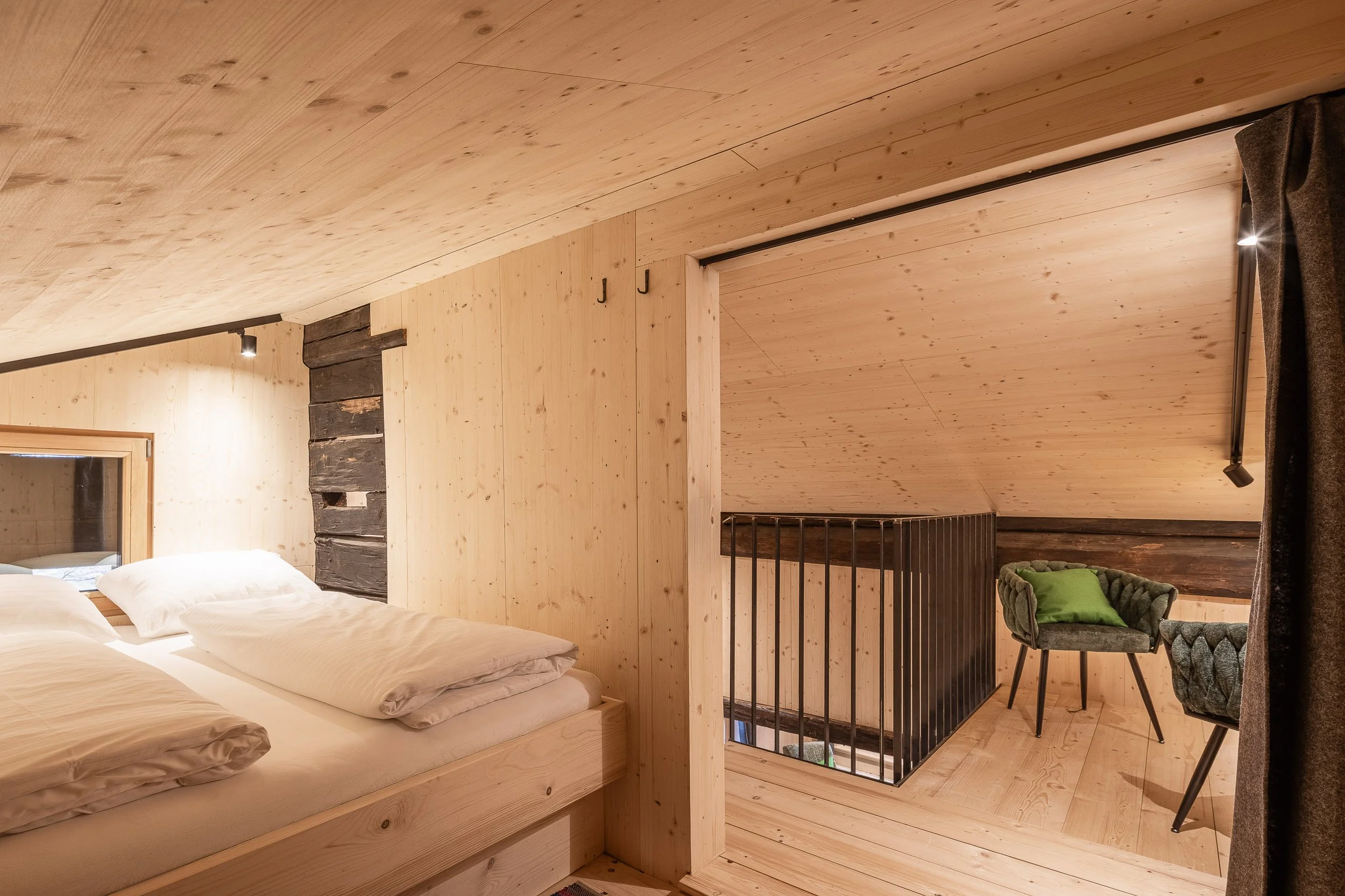 Ein Schlafzimmer mit Holzverkleidung an Decken und Wänden, einem Bett mit weißen Bettwäsche, einem Fenster, einem Holzgeländer einer Treppe, einem Sessel mit grünem Kissen und einer Stehlampe.