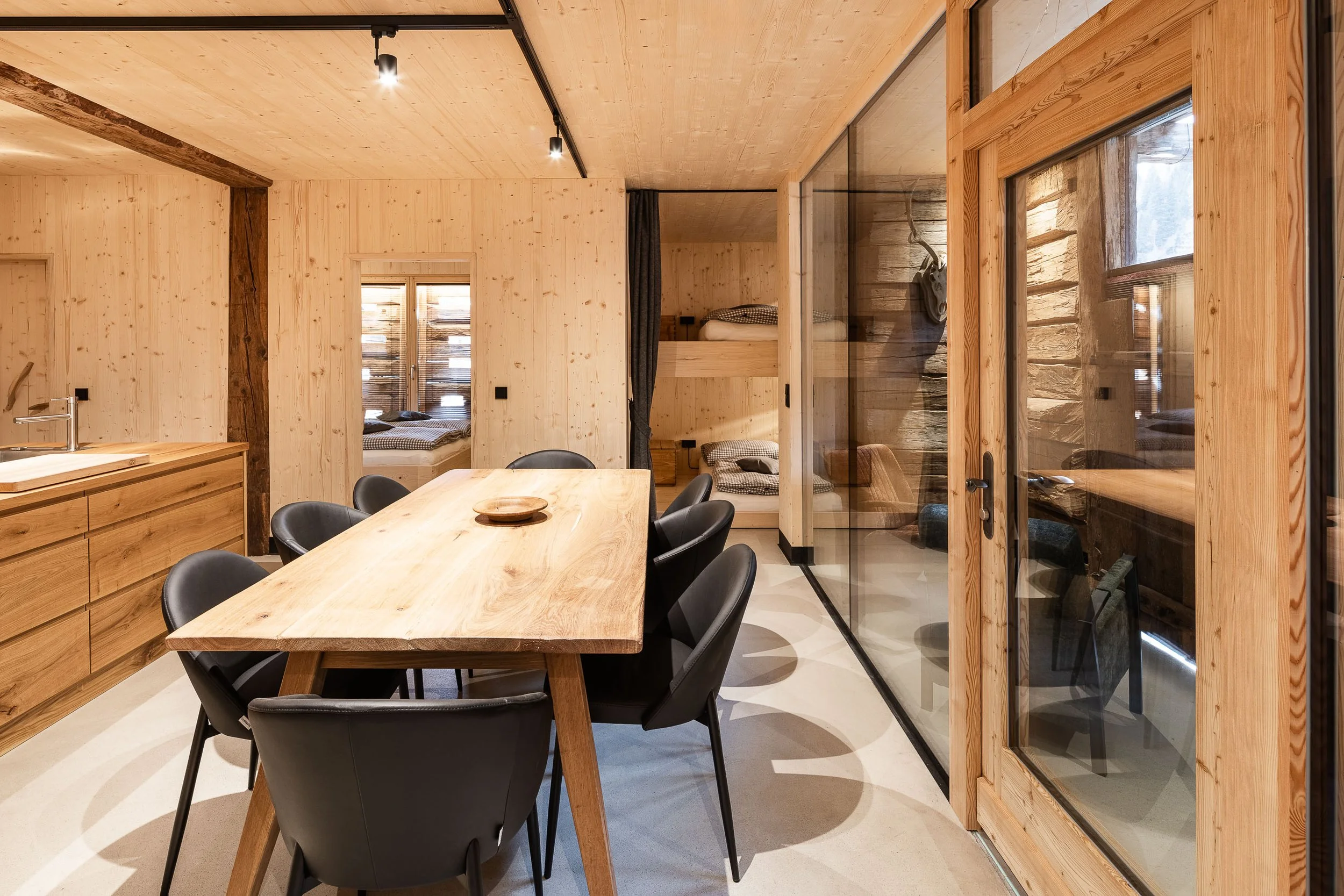 Innenraum eines modernen Hauses mit Holzdekor, Essbereich mit Holz Tisch und schwarzen Stühlen, sichtbar durch eine große Glastür, Schlafzimmer mit Hochbett im Hintergrund, große Fenster, natürliche Beleuchtung.