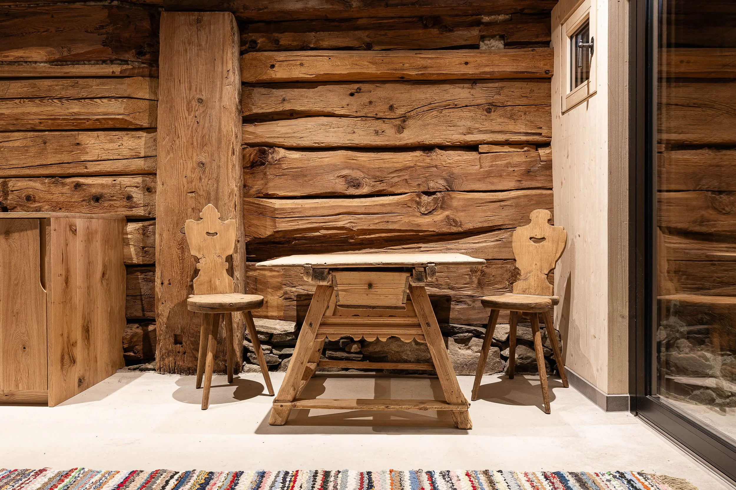 Ein rustikaler Raum mit Holzmöbeln und Holzverkleidung an den Wänden. Es gibt einen kleinen Holztisch, zwei Holzstühle mit geschwungenen Rückenlehnen und einen Teppich im Vordergrund. Das rechte Fenster hat eine geschlossene Holzscheibe, durch die man nach draußen schauen kann.