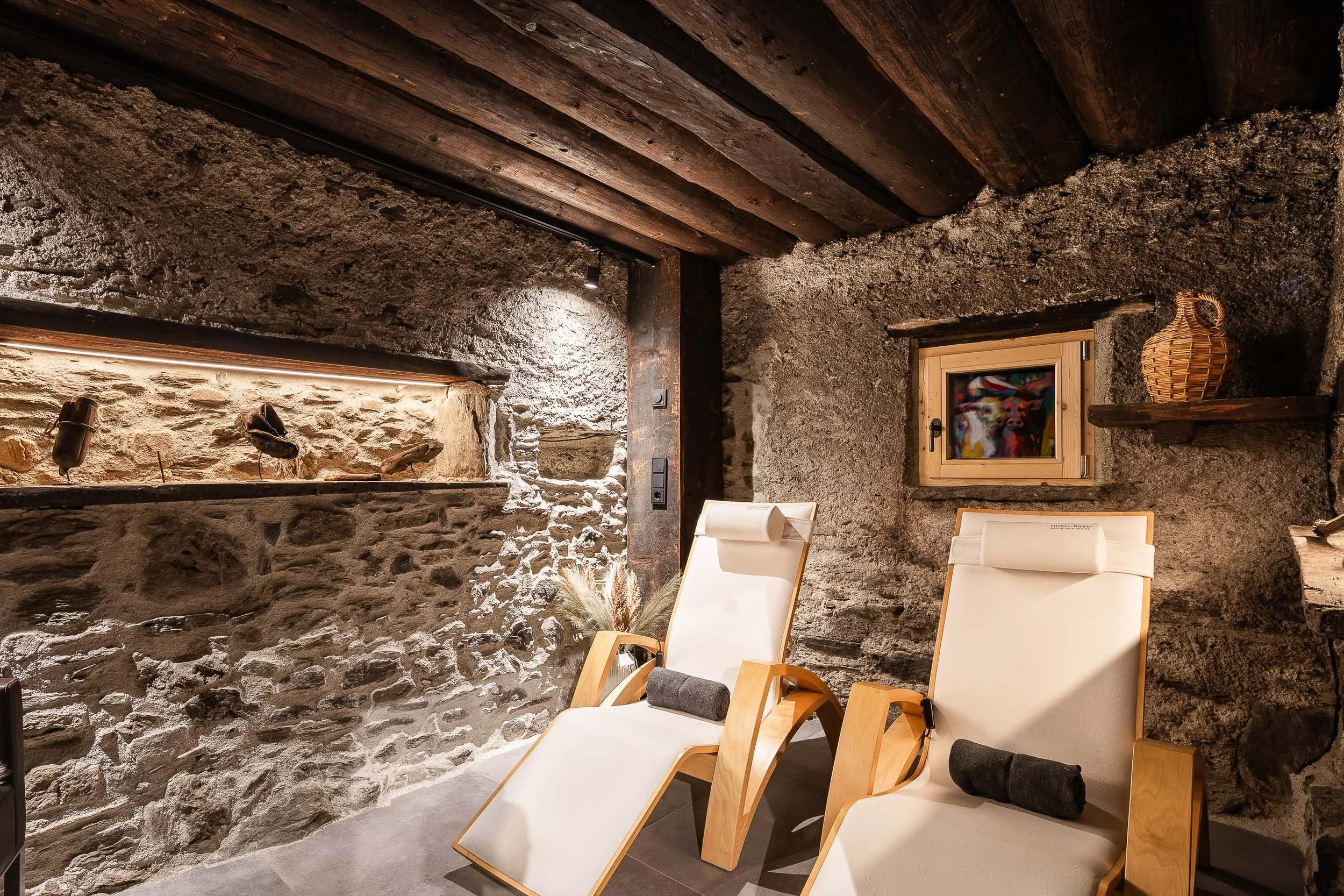 Innenansicht eines rustikalen Zimmers mit Steins- und Holzdecke sowie Steinwänden. Es gibt zwei bequeme Chaiselongues aus hellem Holz mit weißen Matratzen, schwarzen Kissen und Handtüchern. An der Wand befindet sich ein kleines Fenster mit Holzrahmen, ein Korb und eine Wandlampe verleihen dem Raum eine gemütliche Atmosphäre.