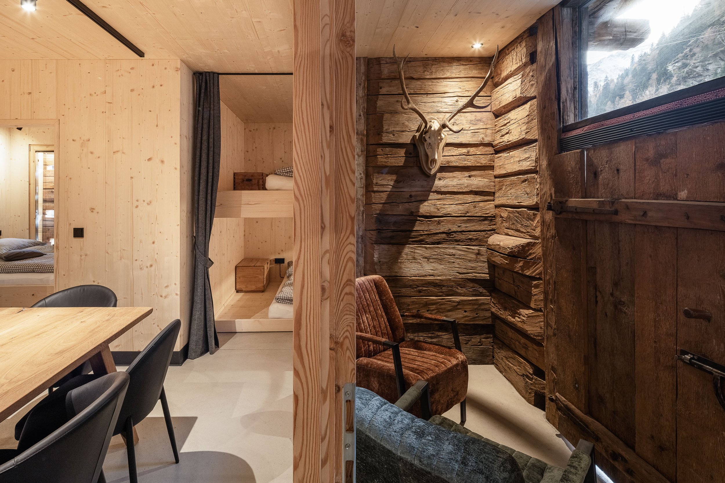 Innenansicht eines Raumes mit Holzverkleidungen, ein Hirschgeweih an einer Holzwand, Stühle und Tische, große Fenster mit Blick auf eine verschneite Berglandschaft.