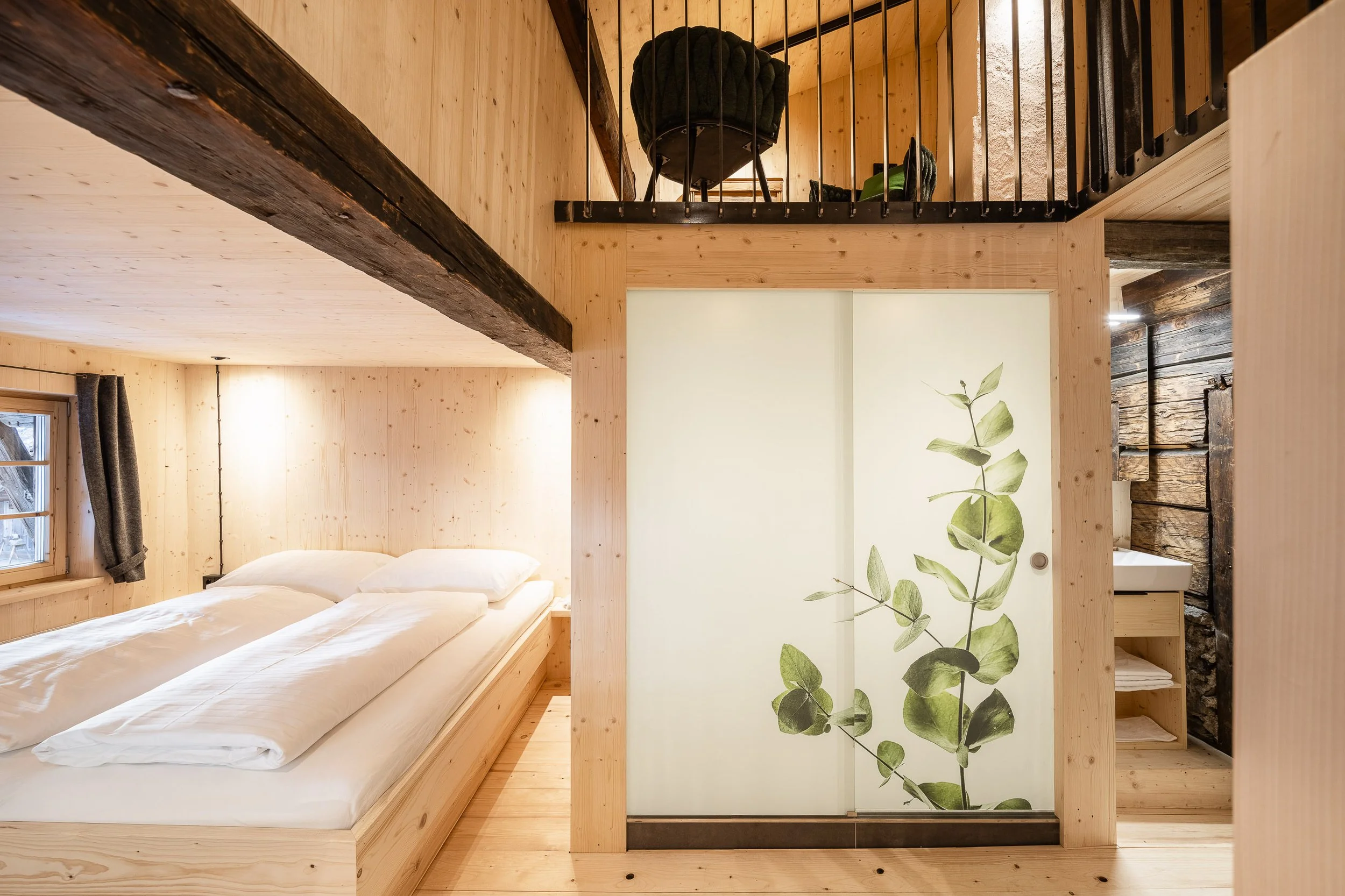 Ein Schlafzimmer mit Holzverkleidung, ein Bett, eine Fenster mit Vorhängen, eine Wand mit einem grünen Blattmuster und eine offene Tür zu einem Badezimmer.