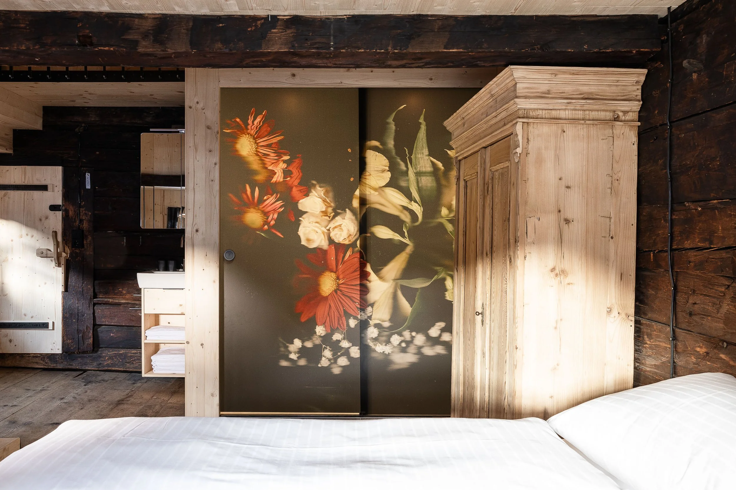 Das Bild zeigt einen Blick in ein Schlafzimmer mit einem Bett im Vordergrund, einem Wandbild mit Blumenmotiven und Holzregalen, eingebettet in eine rustikale Einrichtung aus dunklem und hellem Holz.