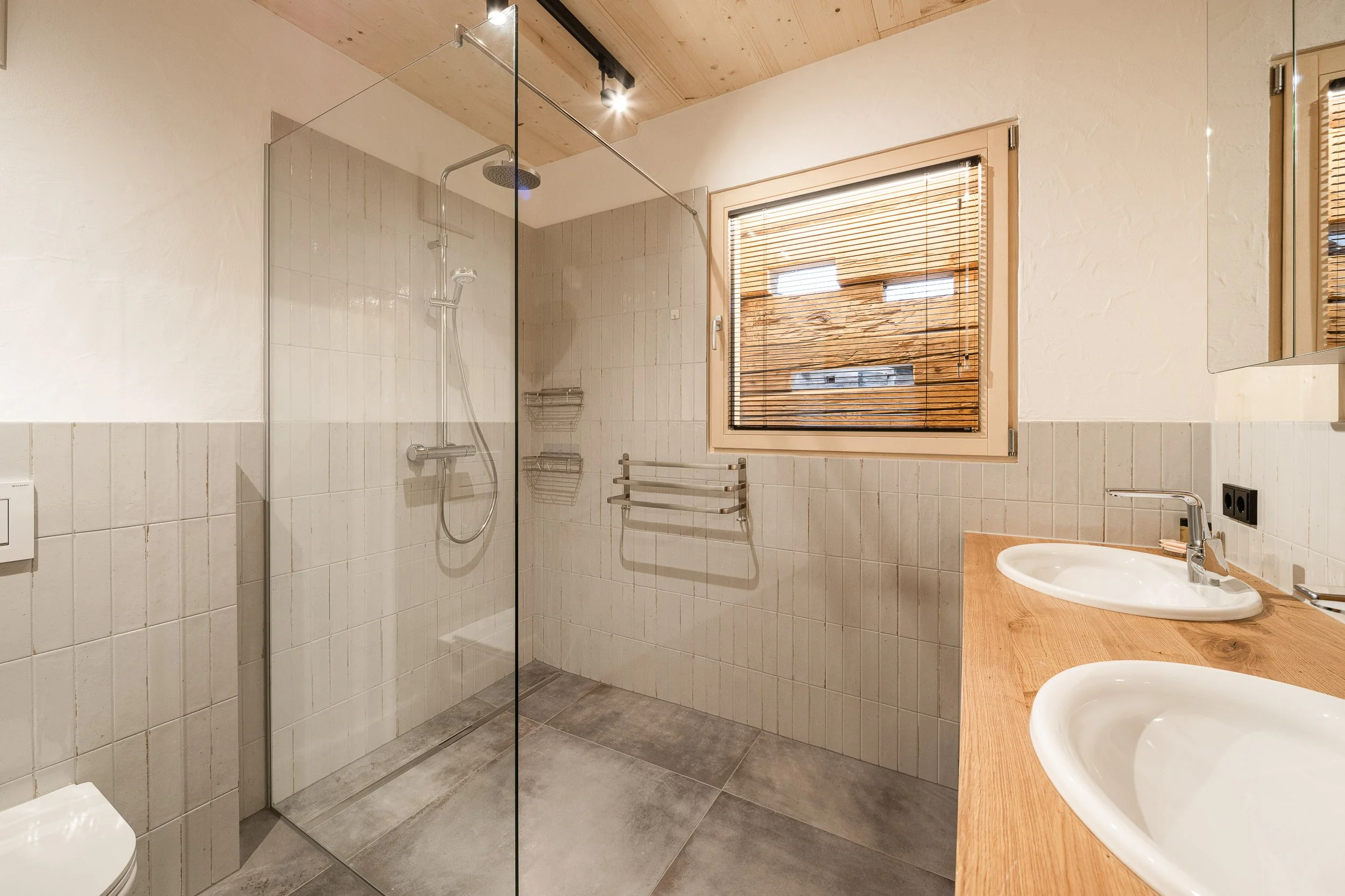 Ein Badezimmer mit einer ebenerdigen Dusche, zwei Waschbecken auf einem Holz-Waschtisch, einer Toilette und einem Fenster mit Holzlamellen. Die Wände sind teilweise gefliest, der Boden ist grau.