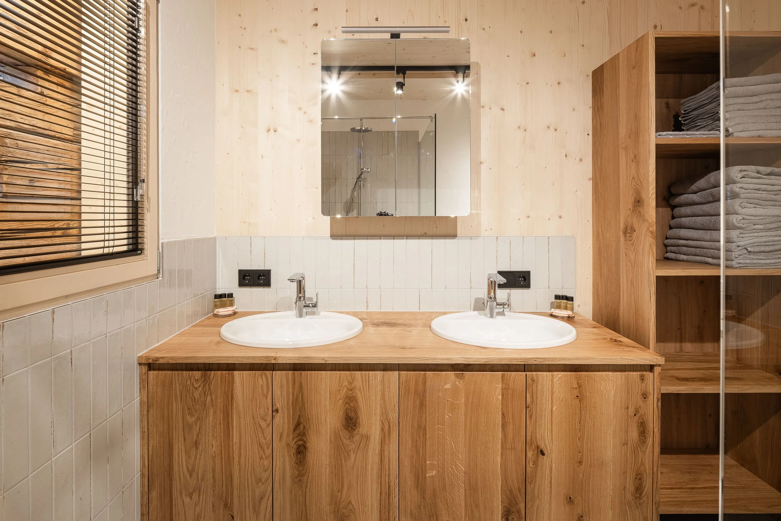 Ein Badezimmer mit Doppelwaschbecken, einem großen Spiegel, Holzregalen mit Handtüchern und einem Blick in die Dusche.