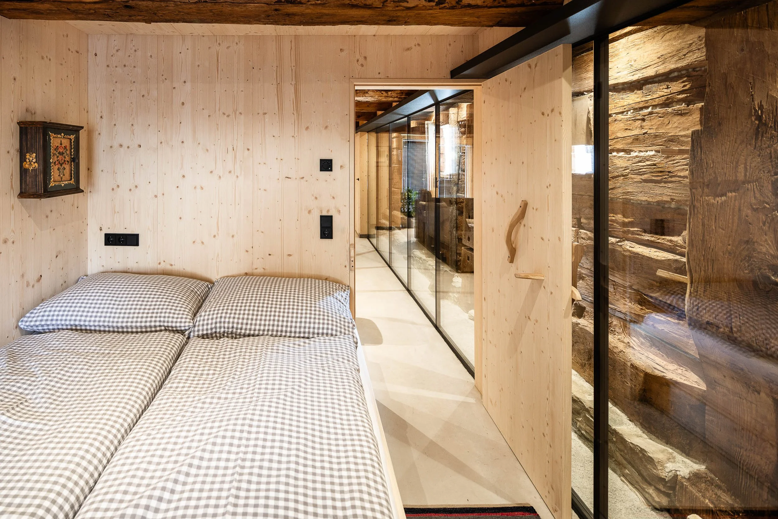 Ein Schlafzimmer mit Holzwänden, ein Bett mit kariertem Bettzeug, eine Trennwand aus Glas mit Holzrahmen und eine Wand aus rohem Holz.