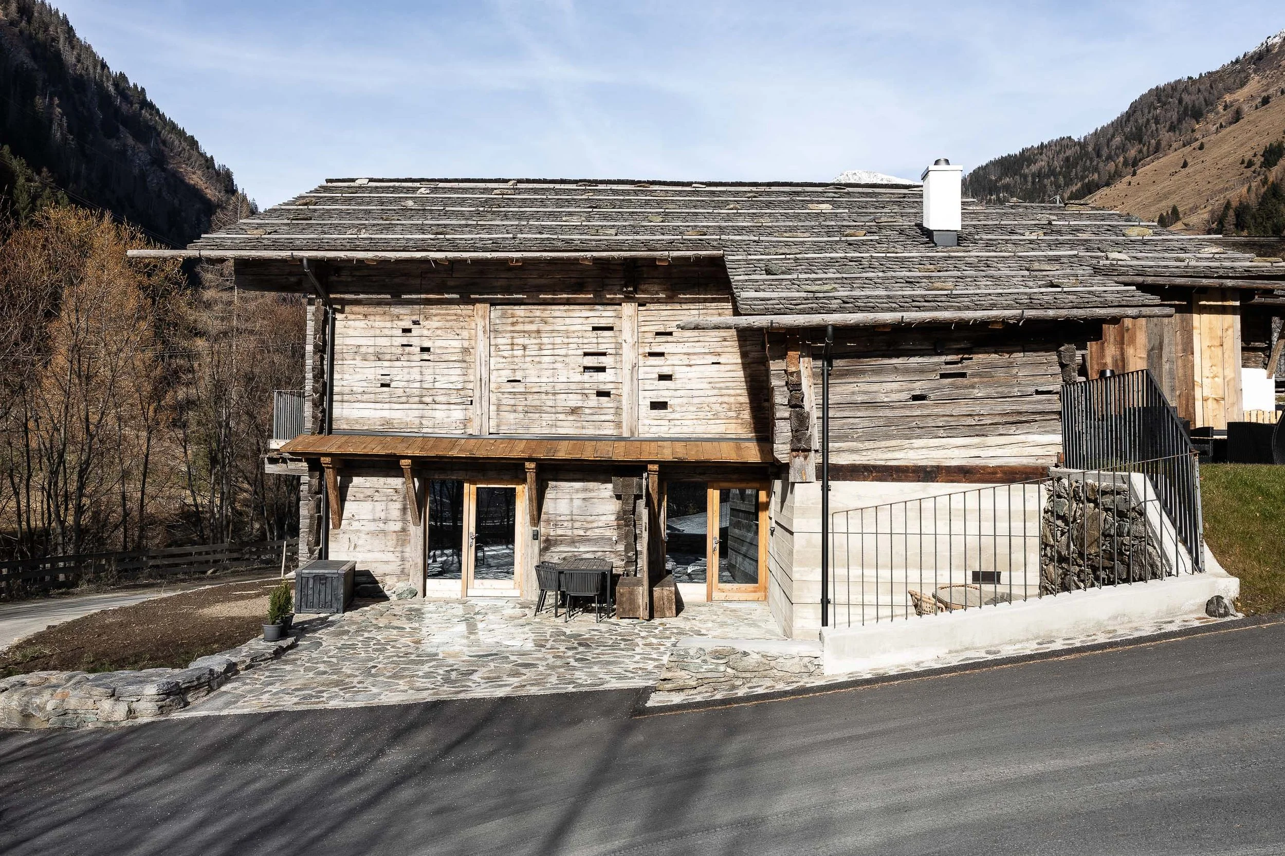 Holzhaus mit Schindeldach, mit Küchen- und Terrassentüren, vor einer Berglandschaft, auf einer Straße in den Bergen.