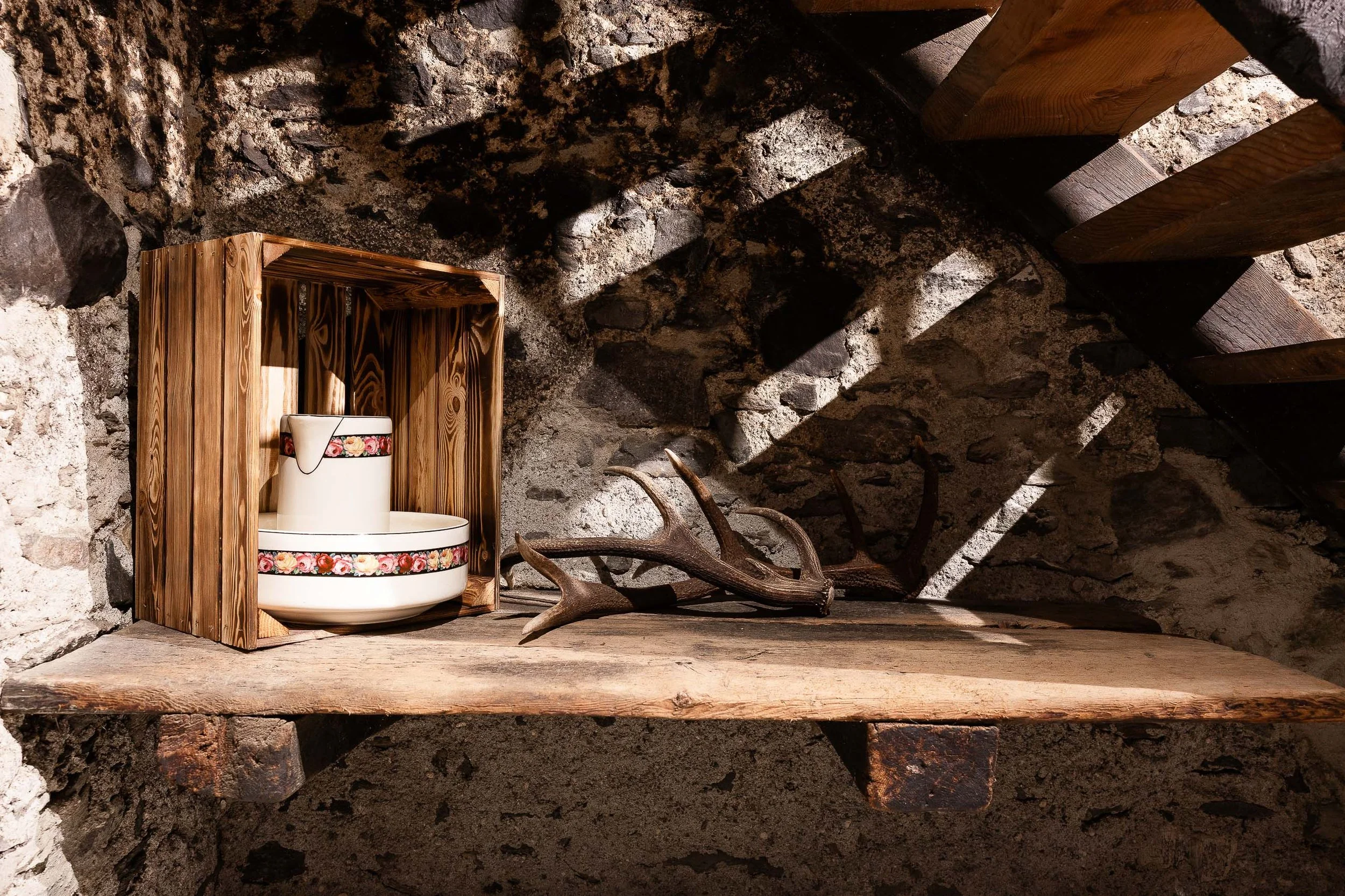 Eine antike Küche mit einem Holzregal, einer weißen keramischen Teekanne mit Blumenmuster, einem hellen Holzboden, einer Steinmauer im Hintergrund und einem Geweih auf dem Boden.