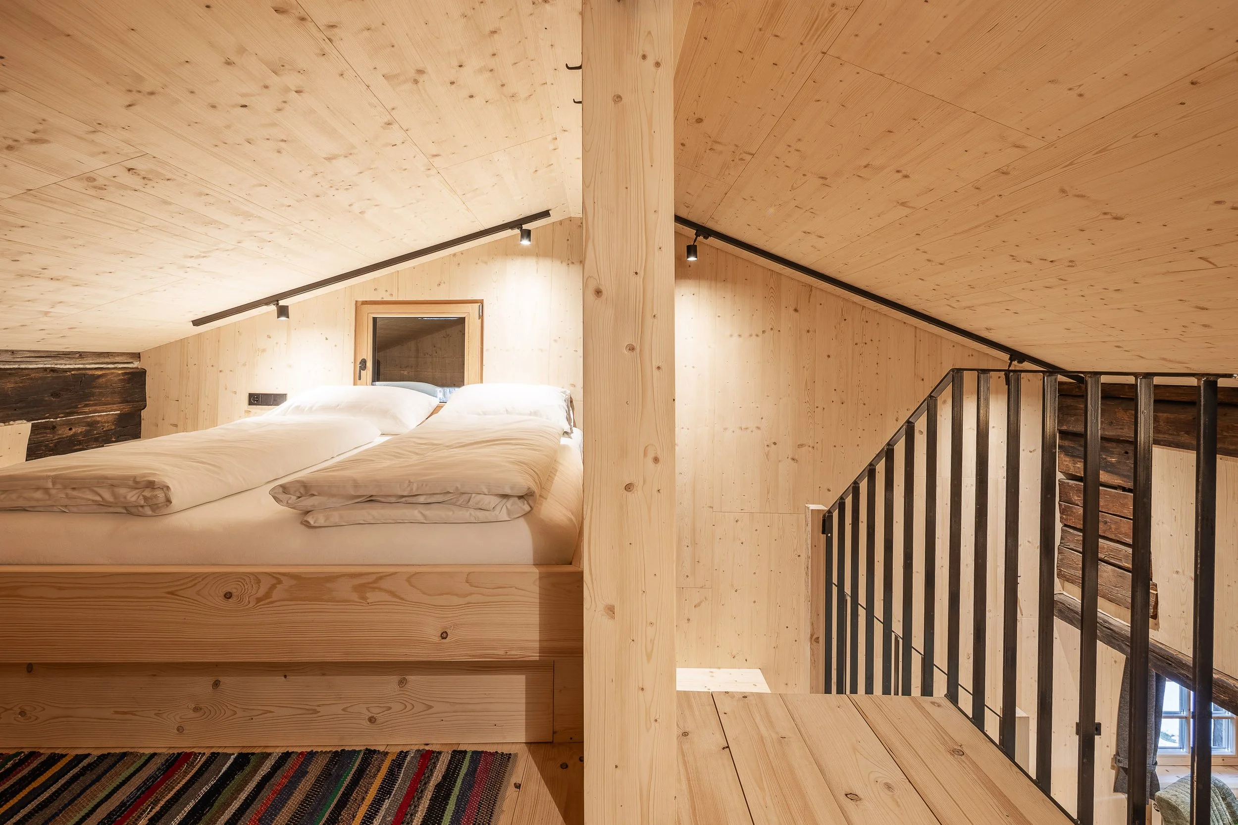 Innenraum eines Zimmers mit Holzwänden und -decken, Doppelbett und Holztreppe.