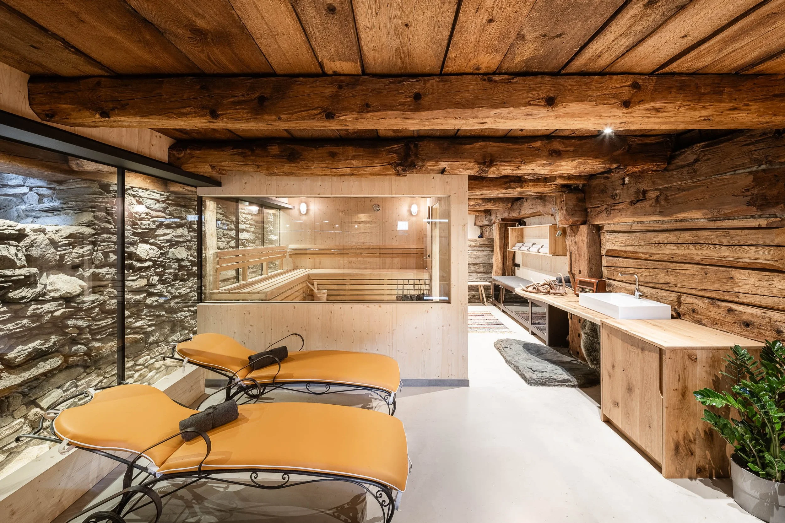 Innenansicht eines Wellnessbereichs mit Holzbalkendecke, Steinfassade, Sonnenliegen und Waschtisch mit Wasserhahn.