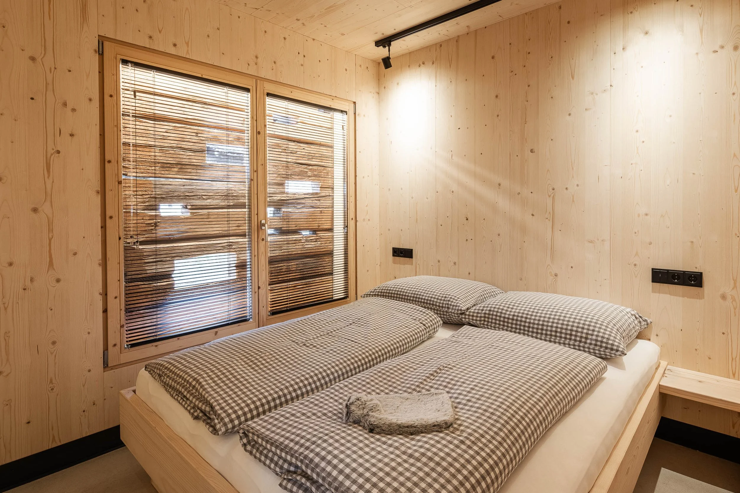 Schlafzimmer mit Holzverkleidung, Doppelbett mit kariertem Bettzeug und Kissen, Fenster mit Holzbalken und Jalousien, Seitentisch mit Felltier