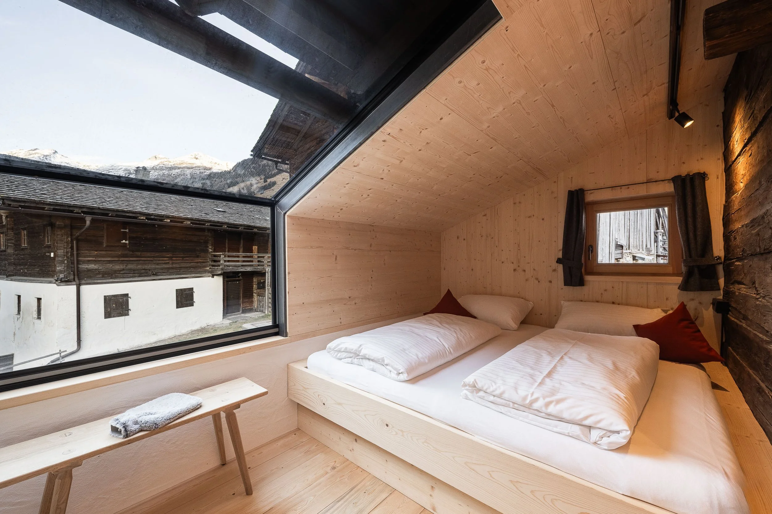 Innenansicht eines modernen Schlafzimmers mit zwei Betten aus hellem Holz, großen Fenstern mit Blick auf schneebedeckte Berge, Holzdecke und -wand, sowie einem kleinen Fenster und dunklen Vorhängen.