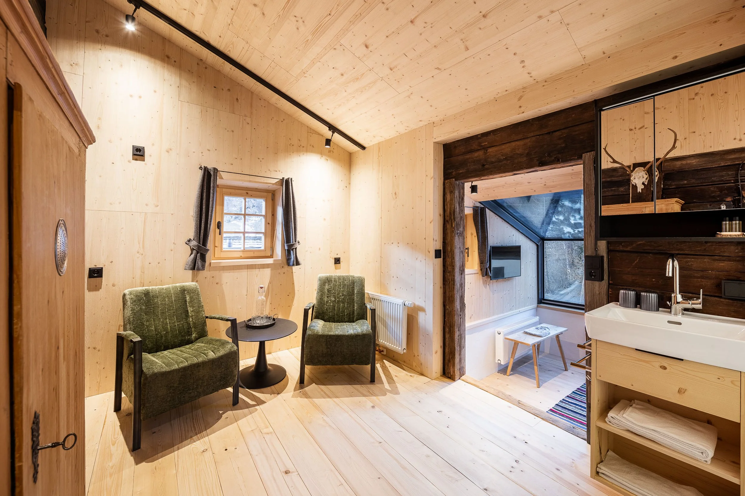 Ein Raum mit Holzwand und -decke, grüne Sesseln um einen kleinen Tisch, Fenster mit grauen Vorhängen, teilweise sichtbare Küche mit Waschbecken und antlerdeko an der Wand.