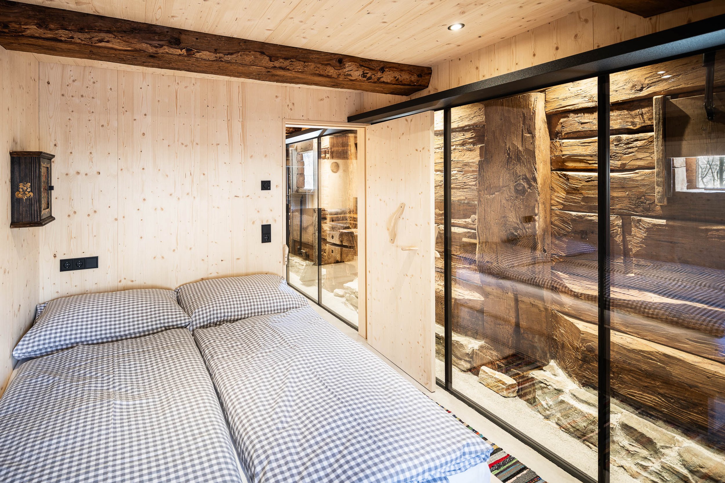 Schlafzimmer mit Holzverkleidung, Betten mit Karo-Bettwäsche, Trennwand aus Holz und Glas, Holzwand mit Fenster, sichtbare Holzbalken an der Decke.