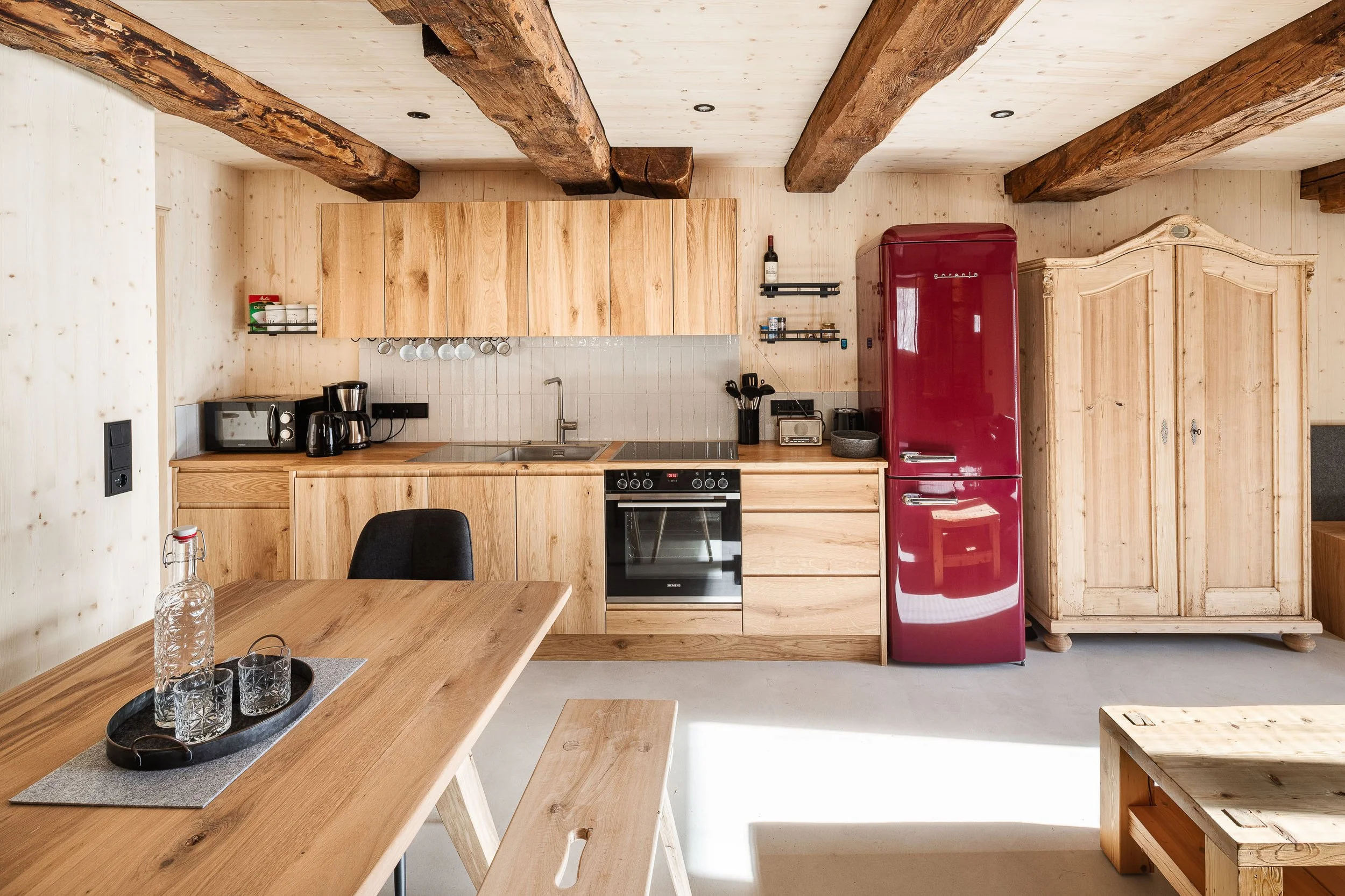 Innenaufnahme einer modernen, rustikalen Küche mit Holzbalken an der Decke, Holzmöbeln, einem roten Kühlschrank, einem Herd und einem Esstisch mit Gläsern.