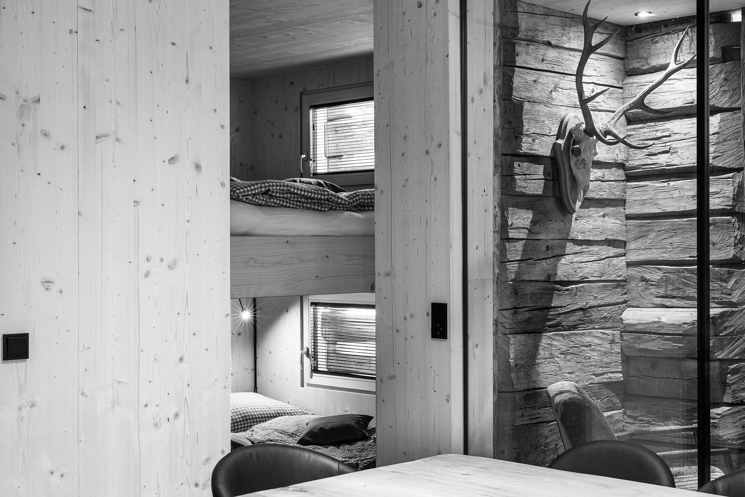 Ein modernes Wohnzimmer mit Holzvertäfelung, großen Fenstern mit Jalousien, einem Bett im Hintergrund, einem Esstisch und Stühlen im Vordergrund, sowie einer Wand mit Warn-Dekoration eines Rehkopfes.