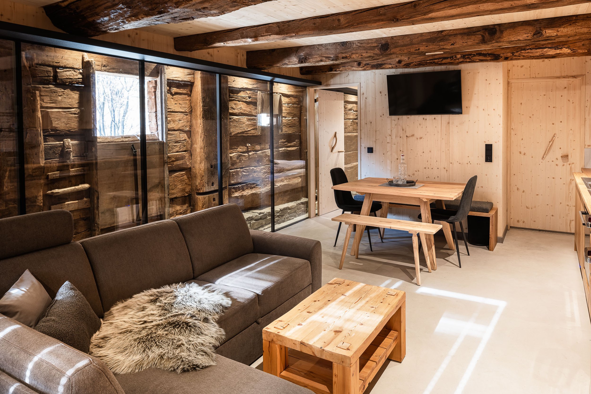 Gemütliches Wohnzimmer mit Holzwänden und -decken, einem braunen Sofa mit Felleinlage, einem rechteckigen Holztisch, einem schwarzen Stuhlkreis, einem Fernseher an der Wand, und einer Glastür, die auf einen winterlichen Ausblick außerhalb zeigt.