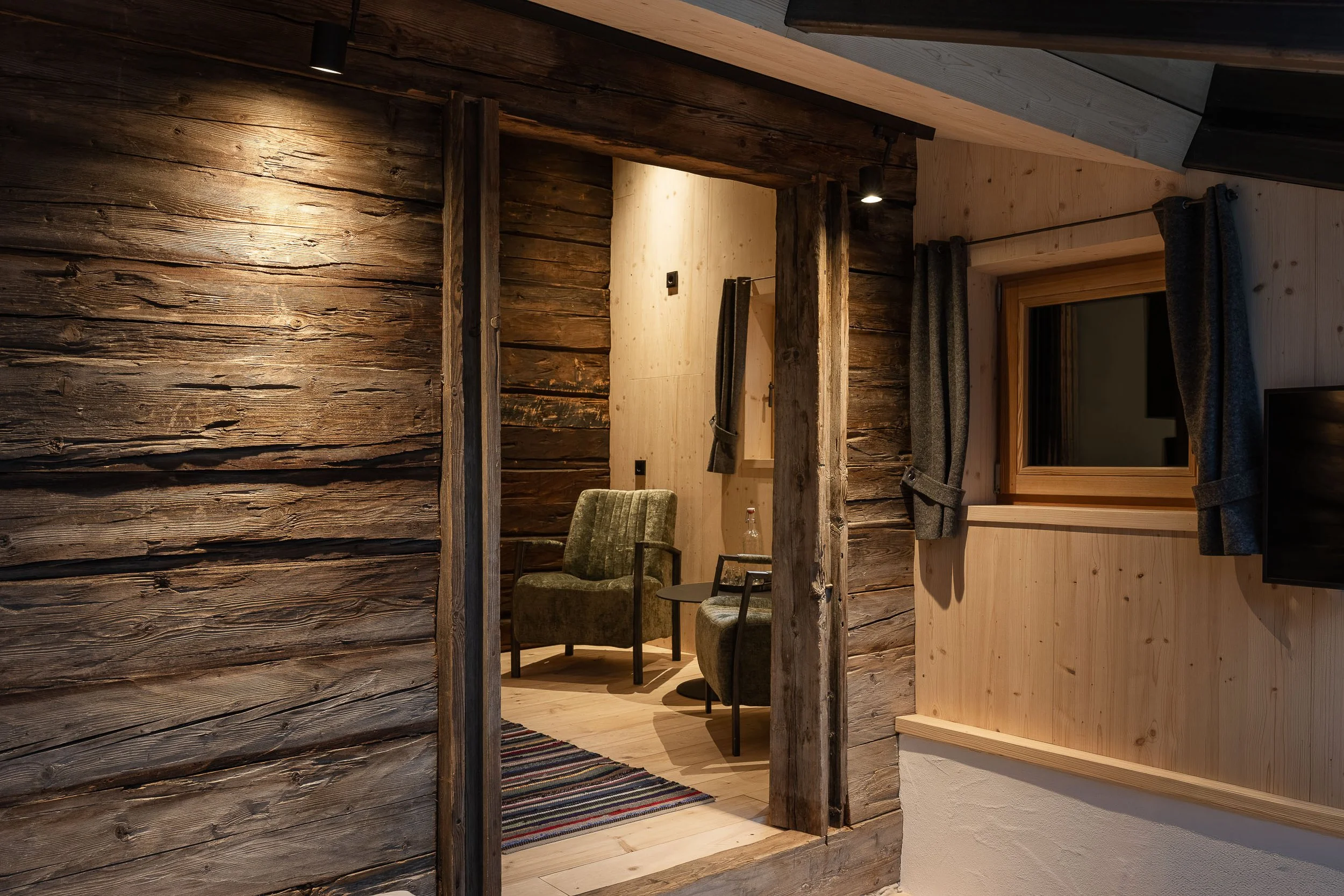 Innenaufnahme eines Raumes mit Holzvertäfelungen, Sessel, einem Teppich und einer kleinen Tafel. Moderne Beleuchtung und eine Holzfensterfront sind sichtbar.