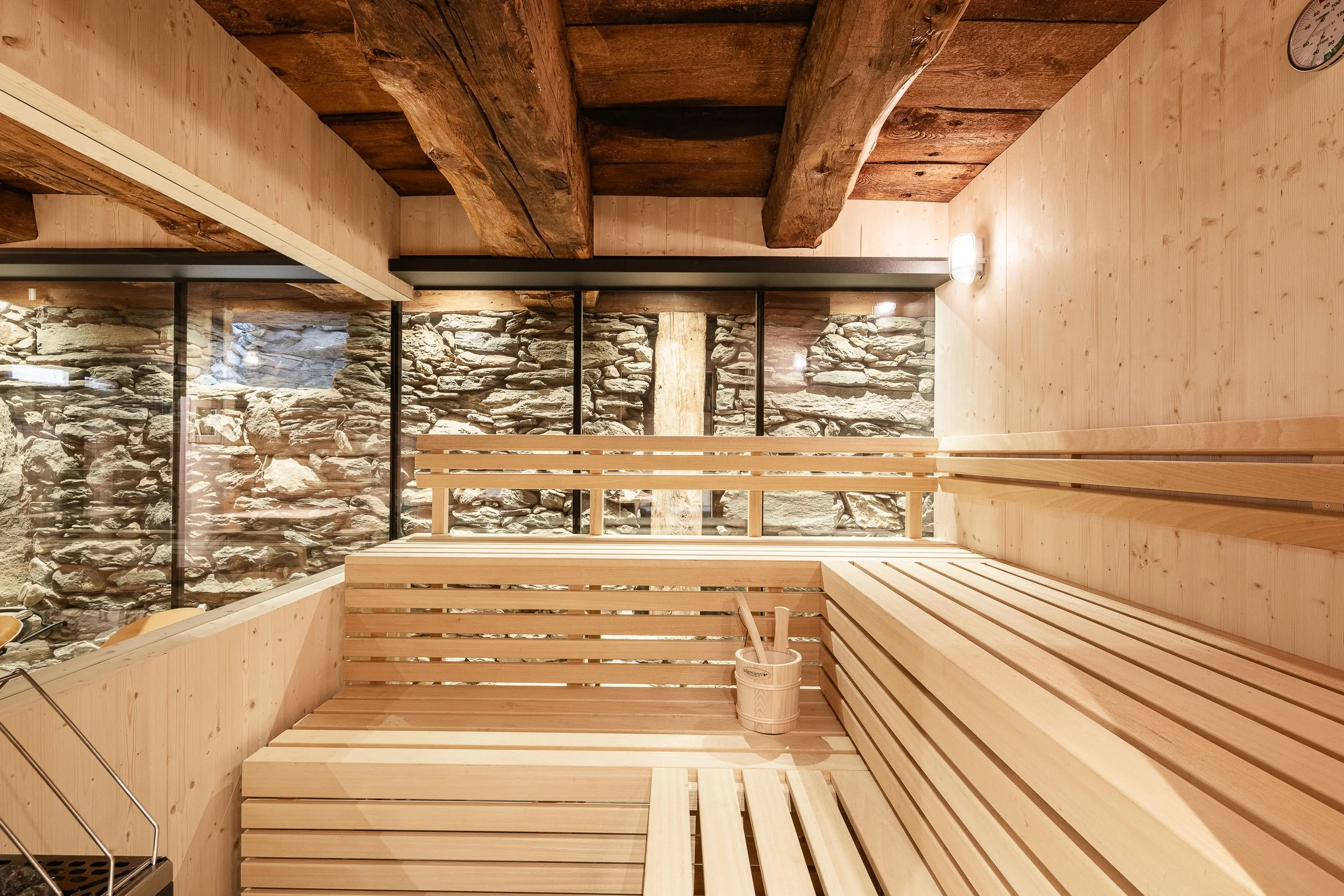 Innenraum einer Sauna mit Holzbänken, Holzwand und Steinwand im Hintergrund, Beleuchtung an der Wand, Sauna-Uhr und ein Eimer mit Butsch.