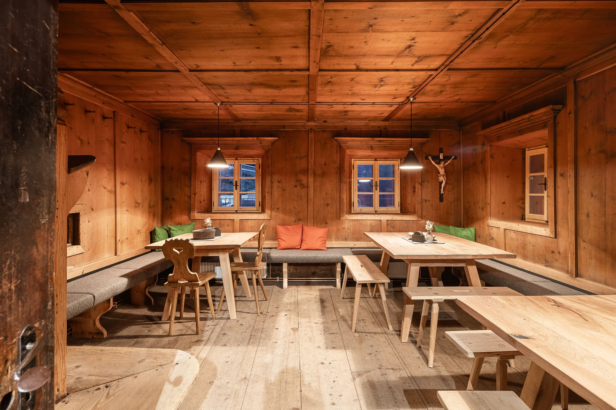 Innenraum eines rustikalen Holzrestaurants mit Holztischen, Holzstühlen, Sofas mit bunten Kissen, zwei Fenster und eine Jesusdarstellung an der Wand.