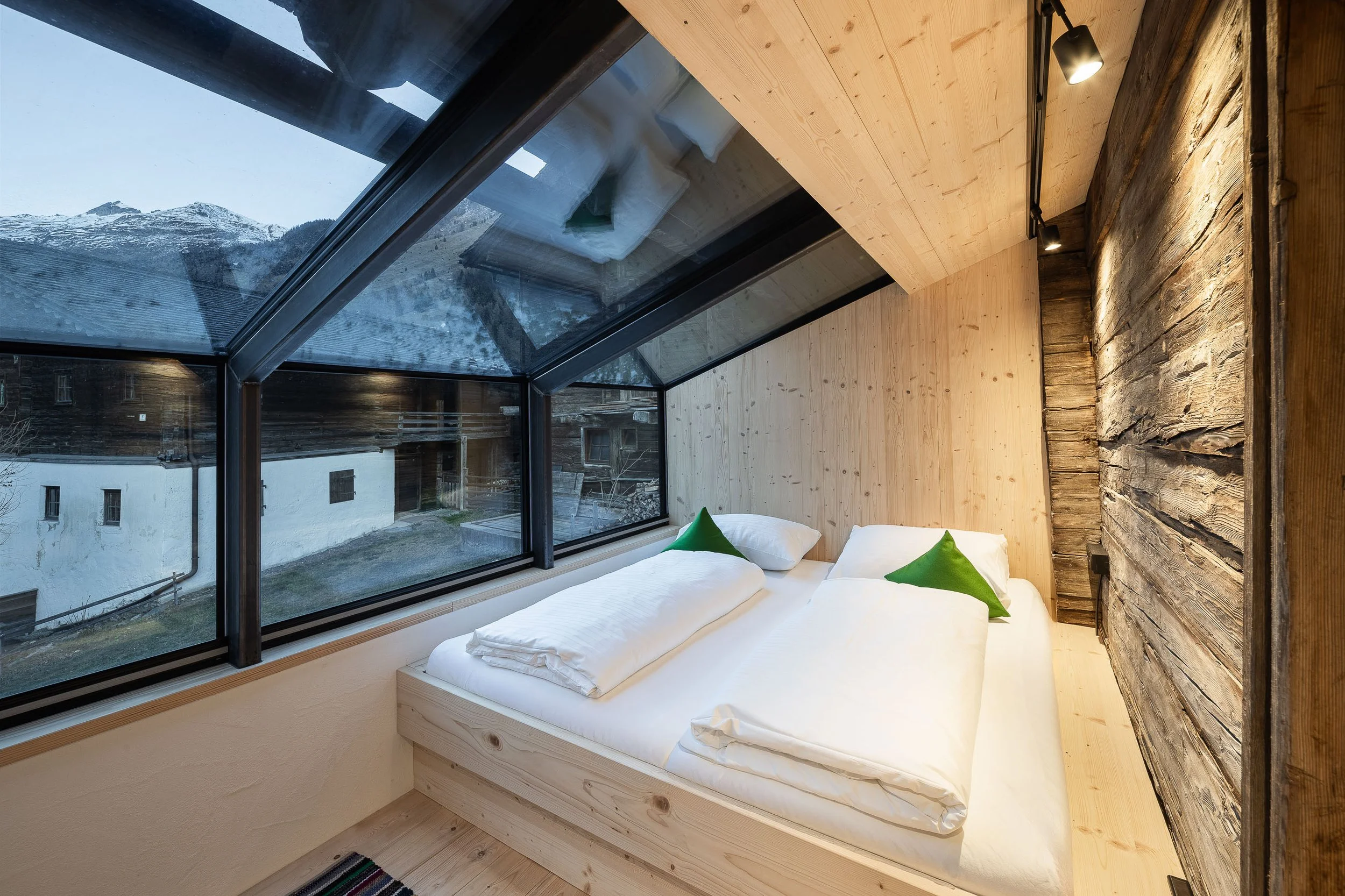 Ein Schlafzimmer mit großen Fenstern, Blick auf Berge, Holzdecke und Wand, Bett mit weißen Bettwäsche und grünen Kissen, moderne Beleuchtung.