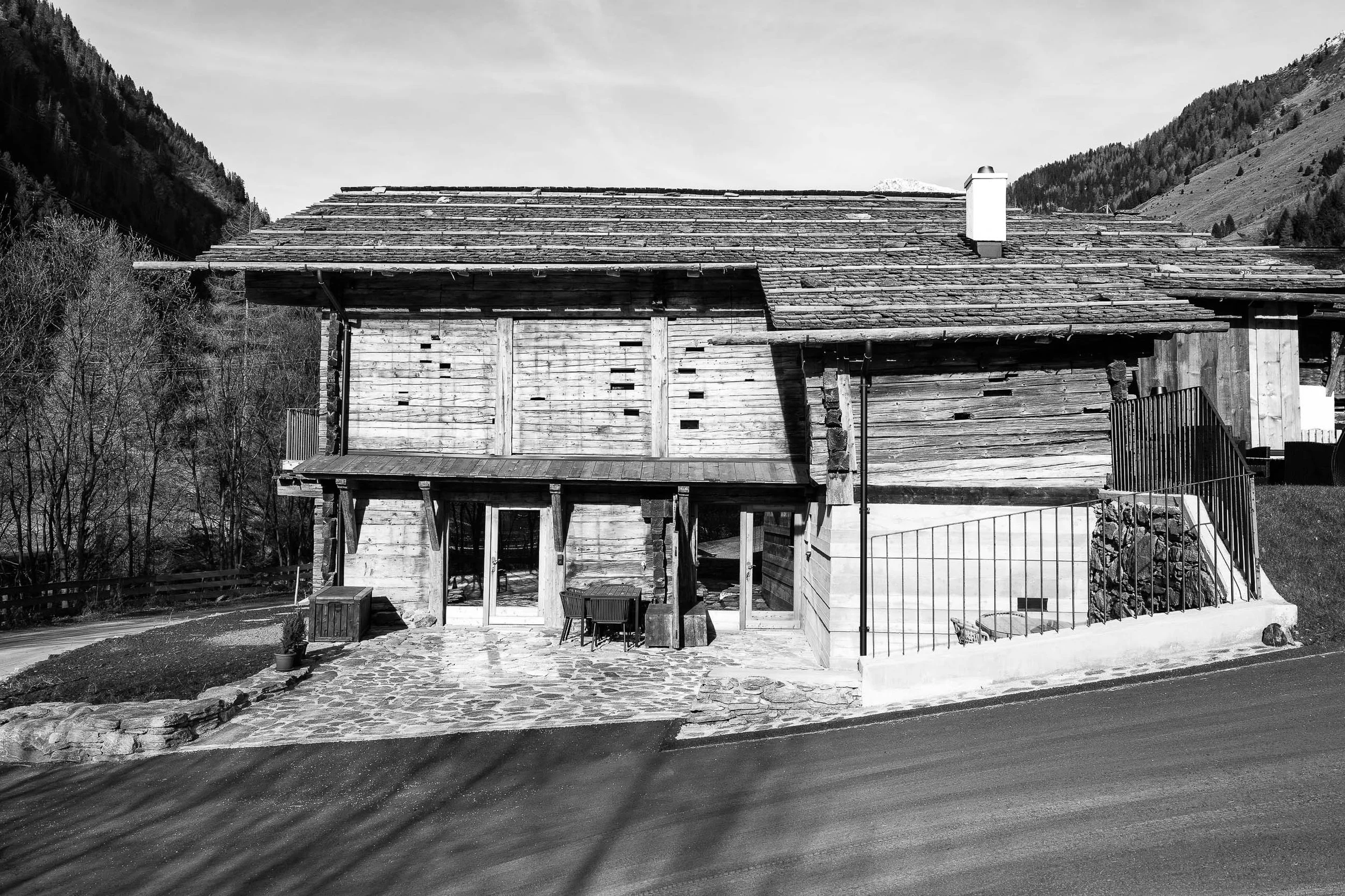 Schwarz-weiß-Foto eines Hauses aus Holz in einer bergigen Umgebung, mit einem Schornstein auf dem Dach, einer kleinen Terrasse mit Möbeln und einer steinernen Einfahrt davor.