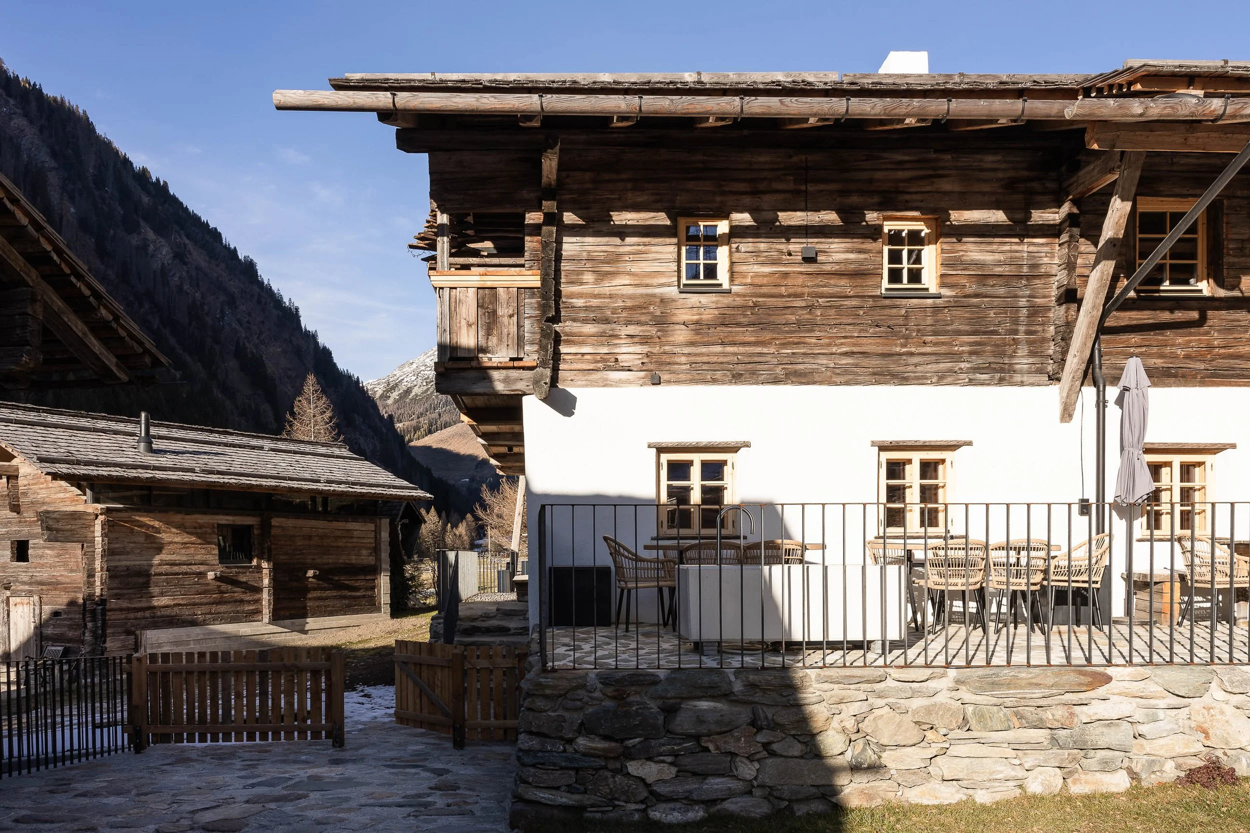 Ein rustikales Holzhaus mit mehreren fenstern, im Hintergrund Berge, ein Tisch mit Stühlen im Außenbereich, umgeben von einer Metallabsperrung.