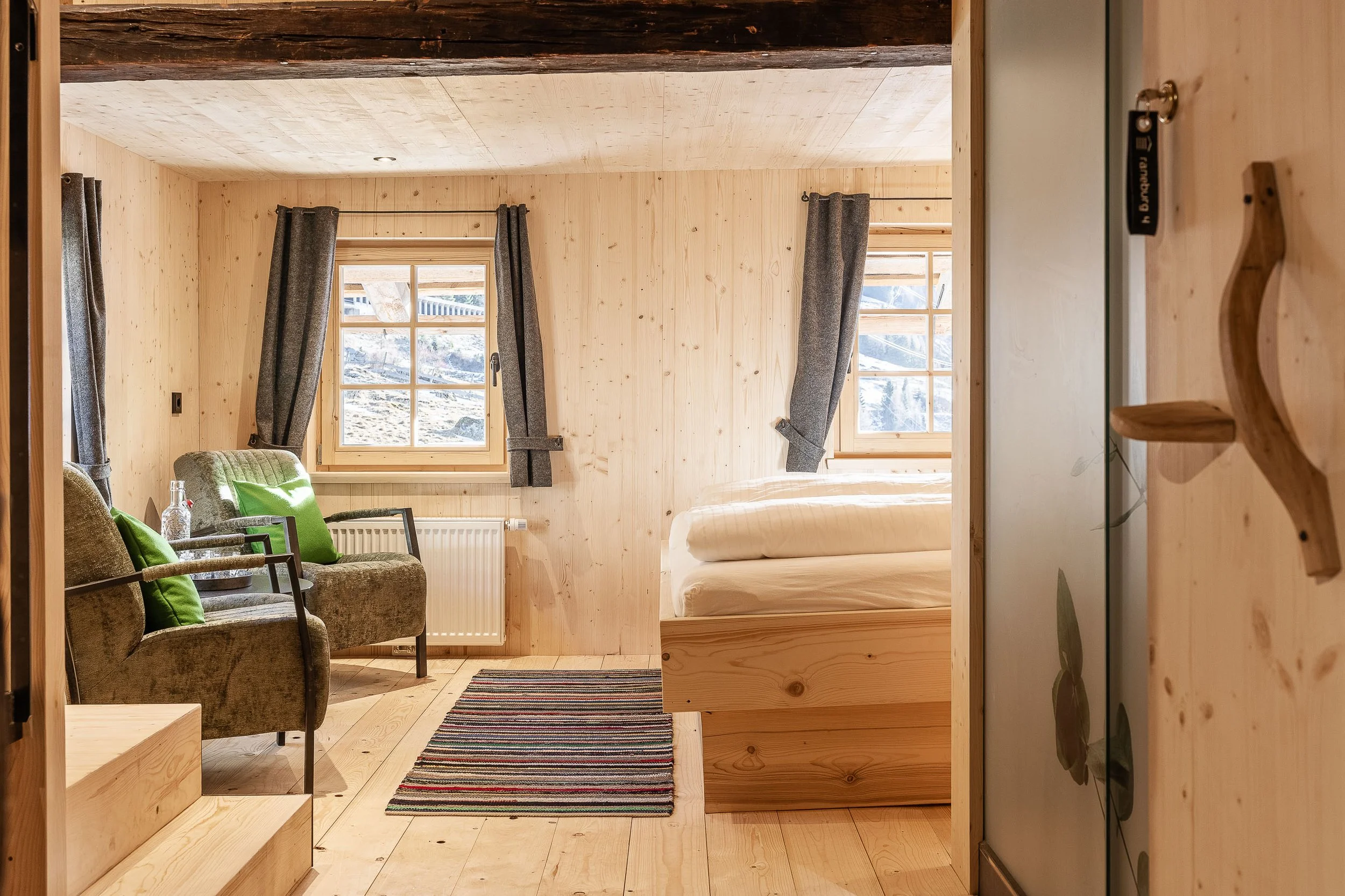 Innenraum eines Holzhauses mit einem Fenster, grauen Vorhängen, einem braunen Sessel mit grünen Kissen, einem kleinen Tisch mit einer Flasche, einem bunten Teppich und einem Bett.