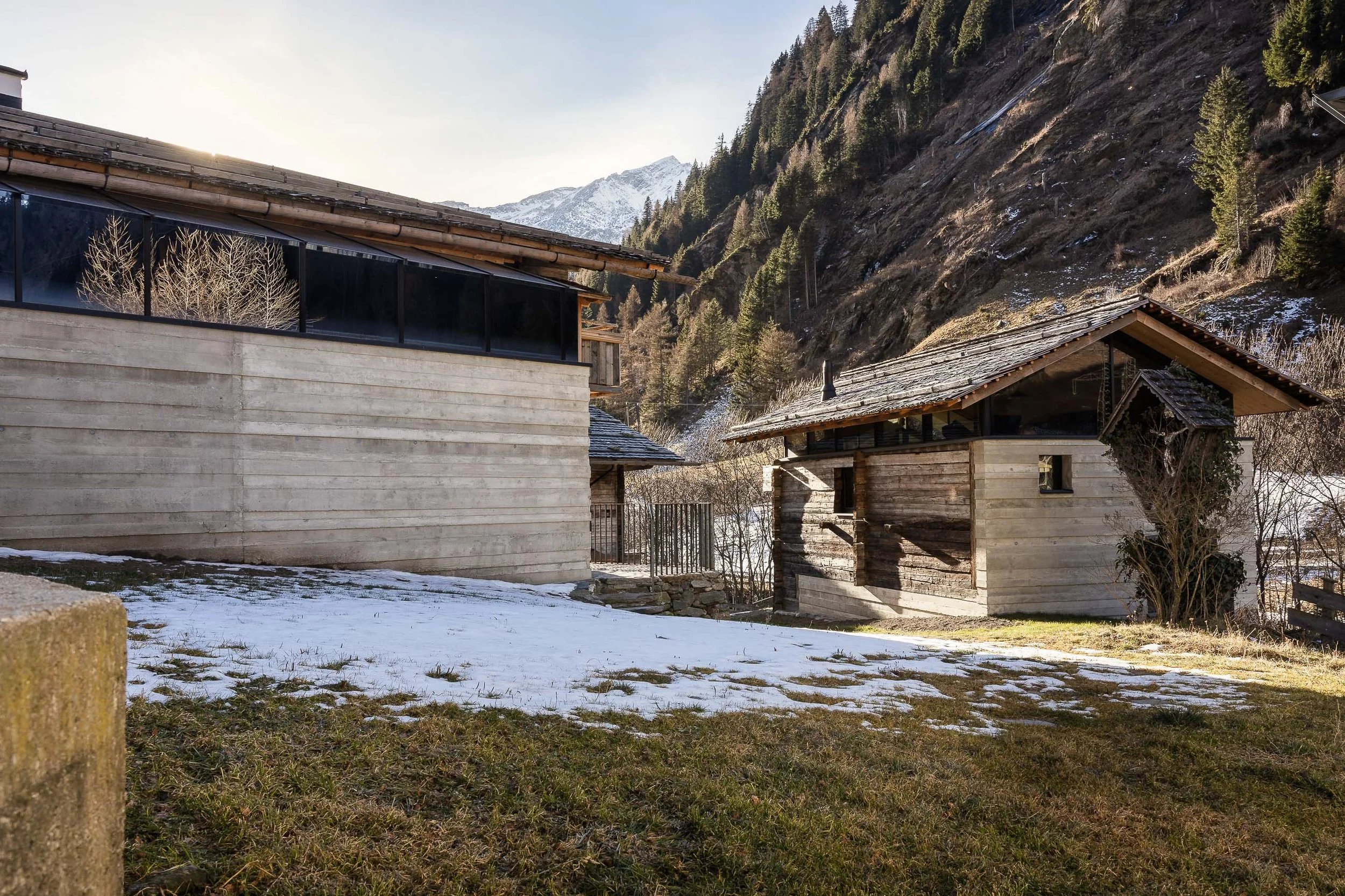 Mehrere Holzhäuser vor einer Bergkulisse mit Tannenbäumen und Schneeresten. Die Sonne scheint im Hintergrund.