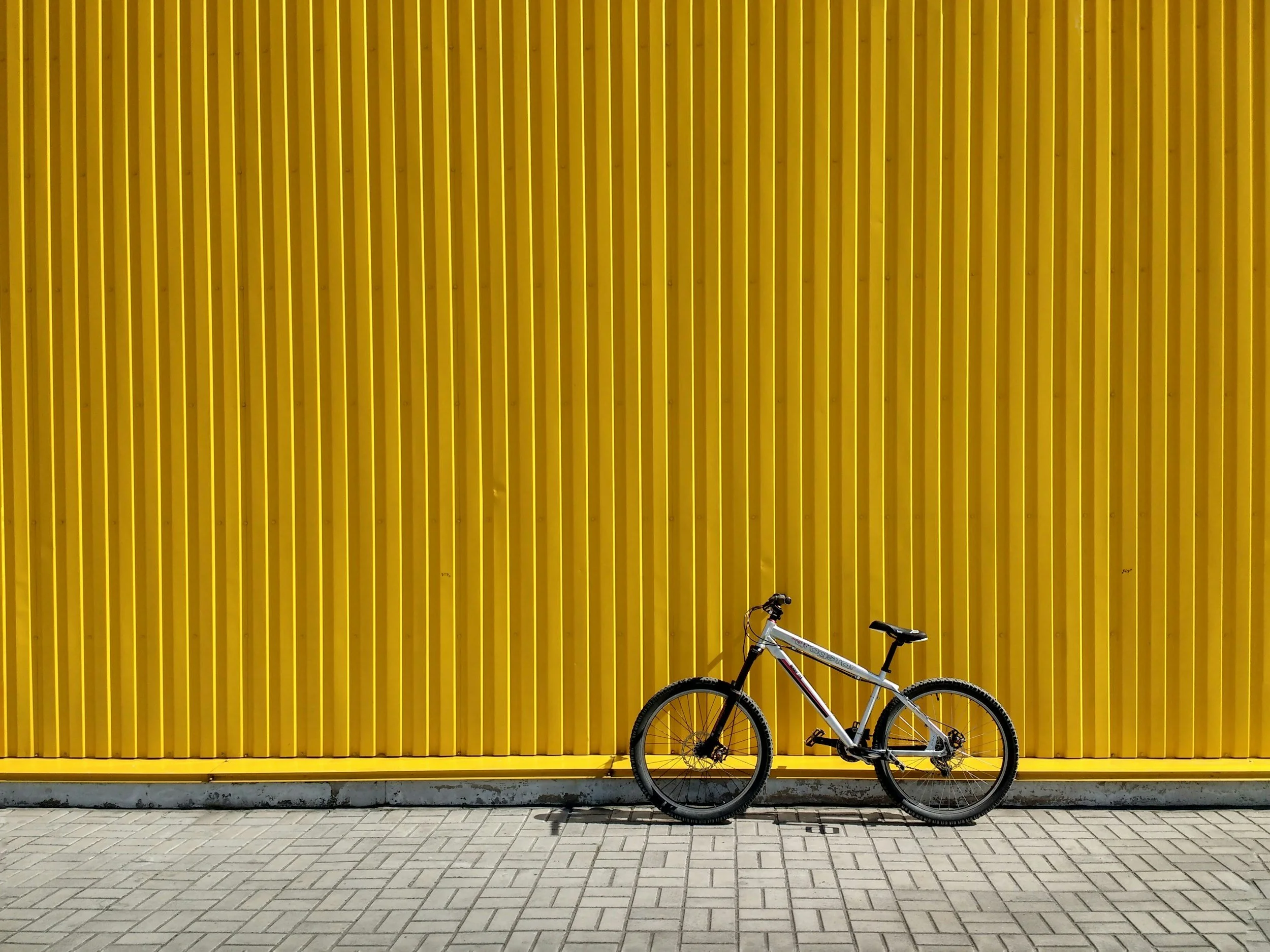A white mountain bike with black tires and a black seat leaning against a large yellow wall with vertical ridges, with a concrete sidewalk in the foreground.
