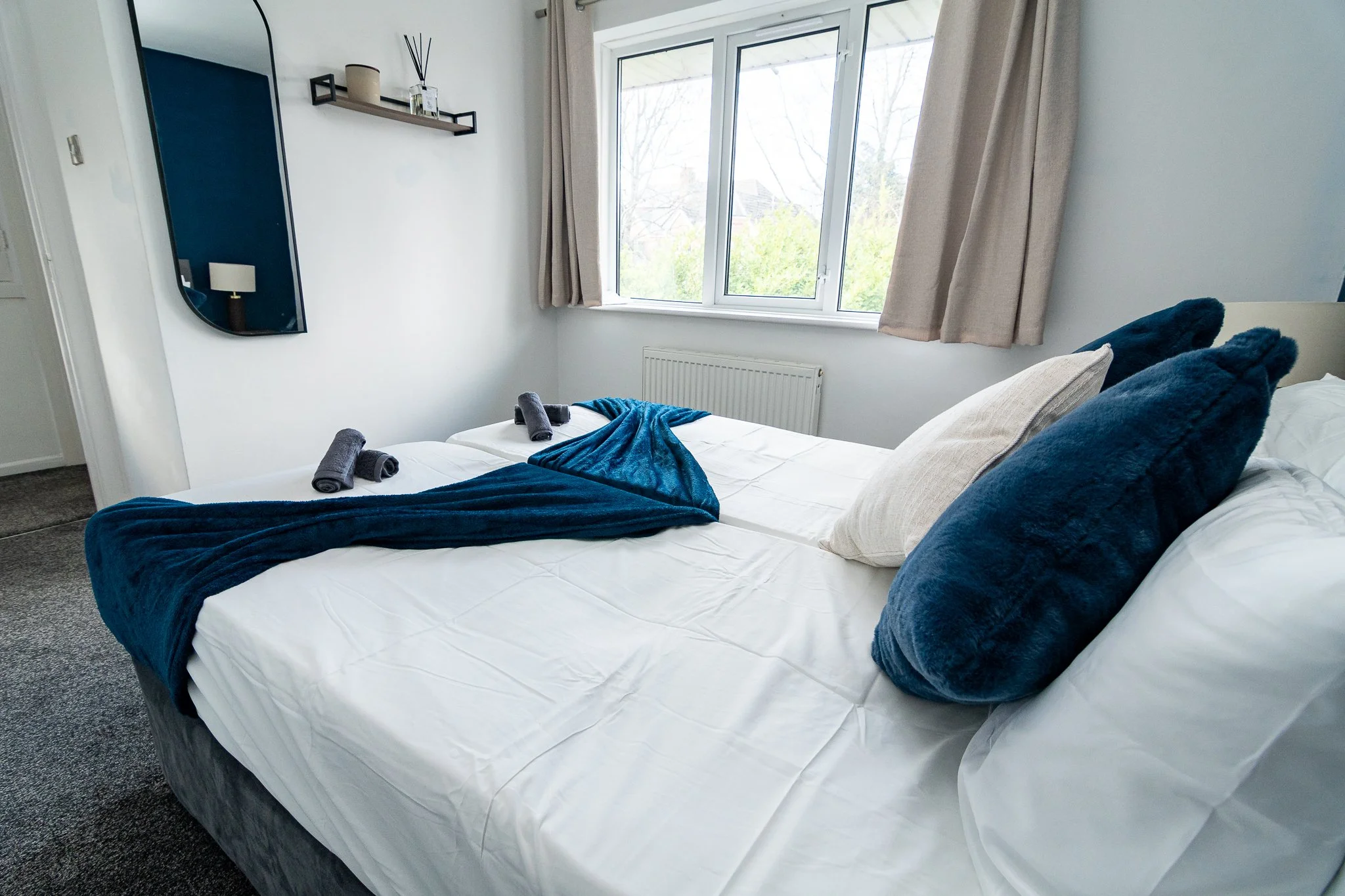 A neatly made bed with white linens, dark blue throw blanket, and matching pillows in a bright bedroom with a large window, beige curtains, and a wall-mounted shelf with decor items.