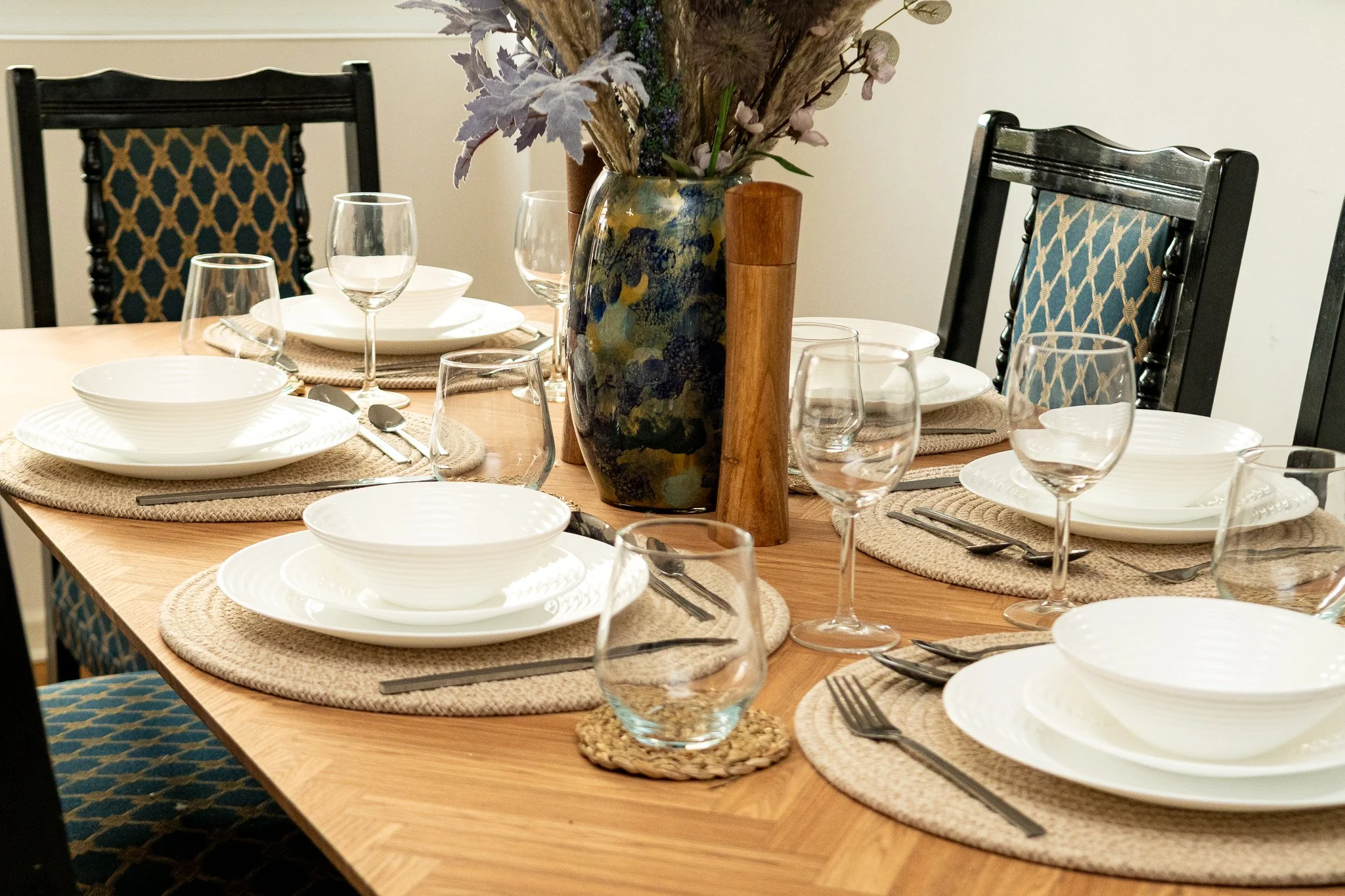 A dining table set with white plates, bowls, and wine glasses on woven placemats. At the center, there is a large floral arrangement with purple and green plants in a colorful vase, accented by a wooden decorative piece. Two chairs with patterned blue and gold upholstery are at the table. The setting appears ready for a meal in a home dining room.