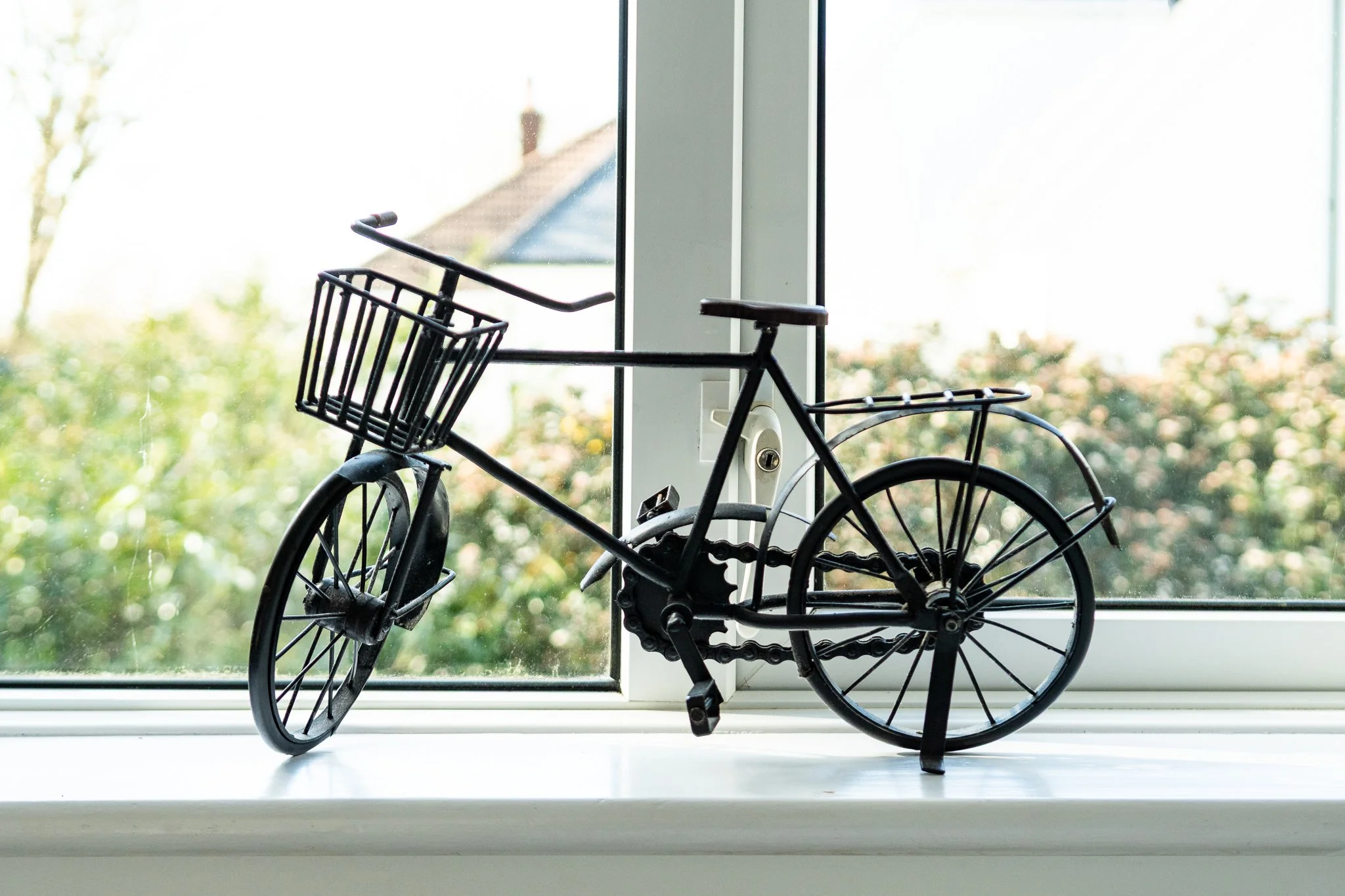 A small black metal bicycle decoration with a front basket, placed on a white windowsill. Behind the window, there is greenery and a house with a sloped roof.