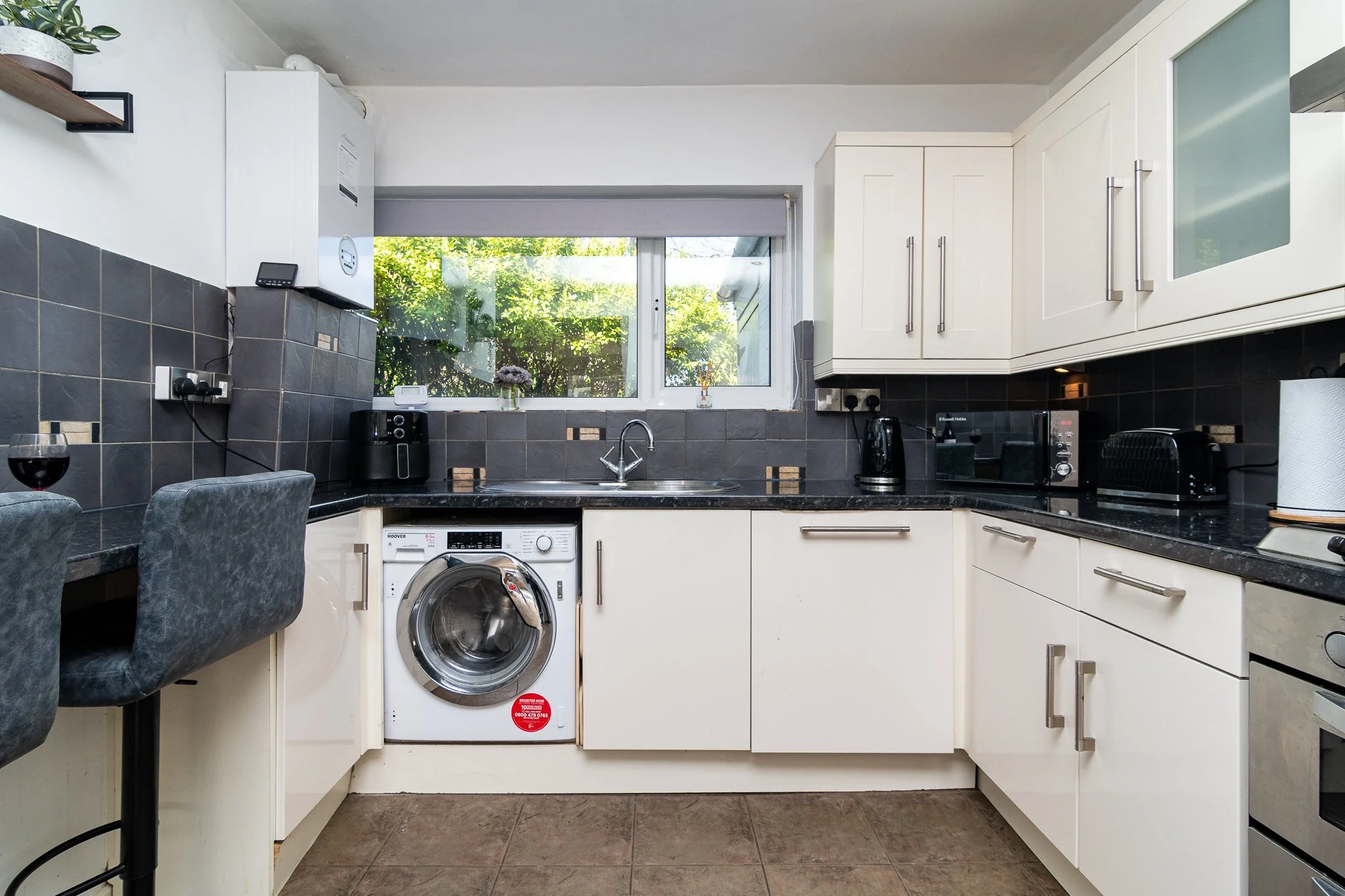 A small kitchen with white cabinets, black countertop, and gray tiled backsplash. There is a window with a view of greenery outside. Appliances include a washing machine, microwave, coffee maker, and toaster. A glass of red wine is on the counter.